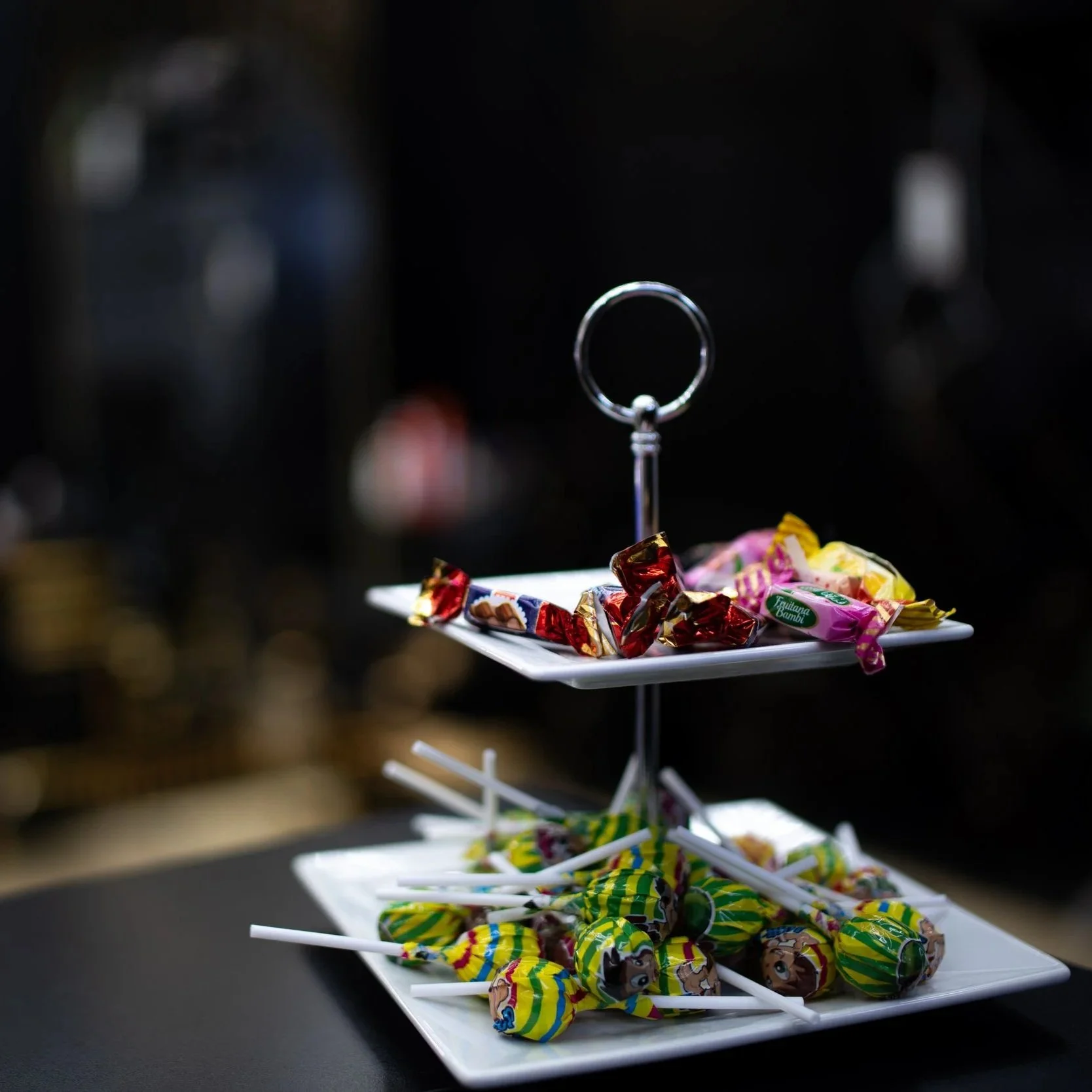 Zwei weiße Teller mit Süßigkeiten, einer auf einer erhöhten Ständerplatte mit gefüllten Lutschern, der andere mit eingewickelten Bonbons, die auf einem schwarzen Tisch stehen.