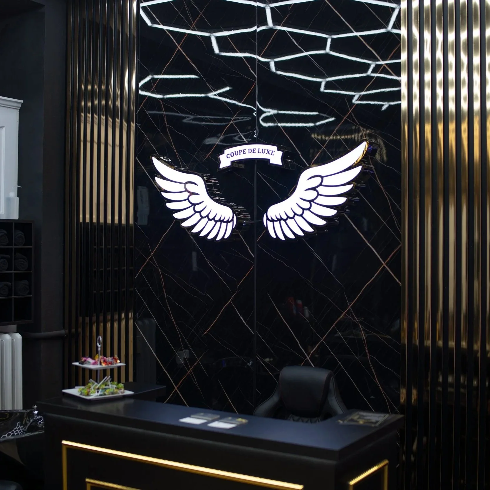 Empfangstresen mit einem schwarzen Schreibtisch, auf dem eine zweistöckige Snackplatte mit verschiedenen kleinen Speisen steht, und ein schwarzer Chefsessel. Hinter dem Tisch an einer schwarzen Wand mit goldenen Linien befindet sich ein Neon-Schild mit Flügeln und dem Schriftzug "Coupe de Luxe".