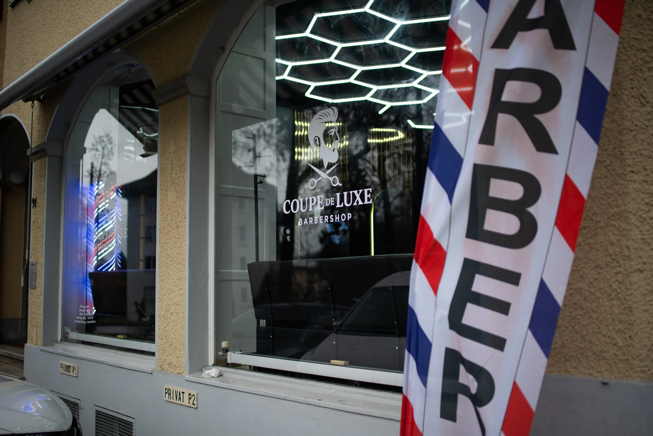 Außenansicht eines Friseursalons mit einem großen Fenster, in dem ein Logo mit einem Haarschnitt und Schneideschere zu sehen ist, sowie der Schriftzug 'COUPE DE LUXE BARBERSHOP'. Im Vordergrund hängt ein Straßenbanner mit der Aufschrift 'HAARSCHNITT'