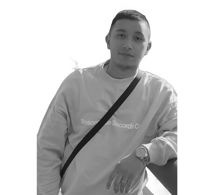 A young man wearing a sweatshirt and a wristwatch, with a shoulder bag, standing against a dark background.