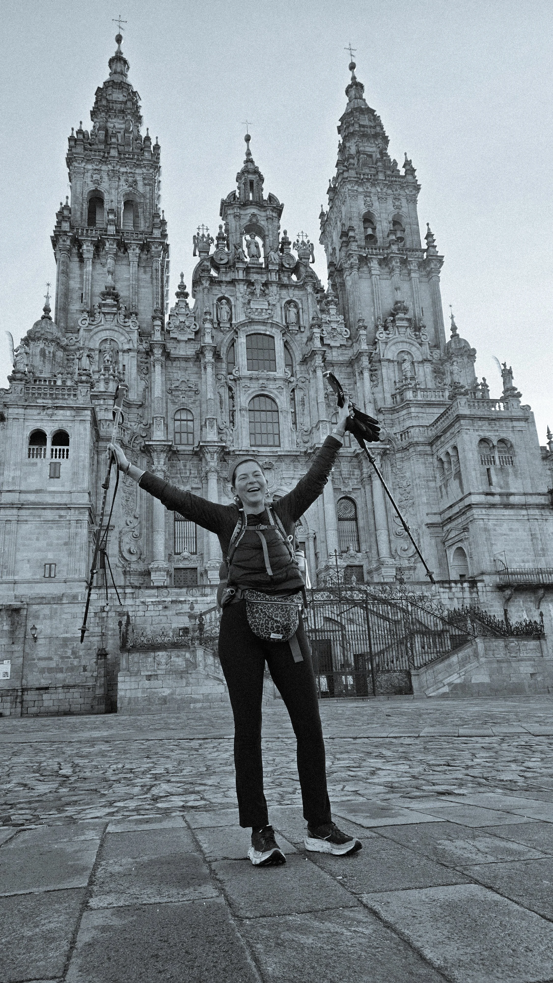 Une femme souriante avec des bras levés en tenant des bâtons de marche devant une grande cathédrale baroque, probablement à Santiago de Compostela.