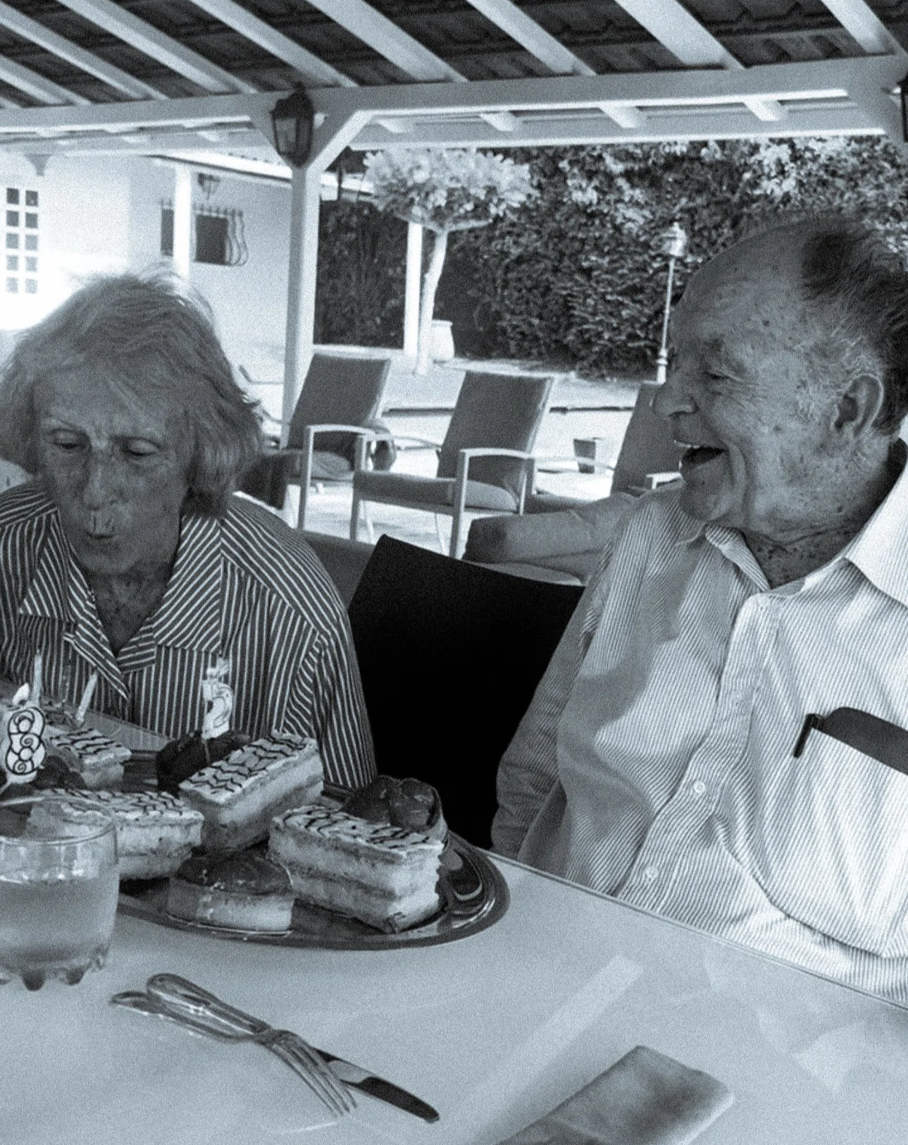 Une photo en noir et blanc de deux personnes âgées qui semblent célébrer un anniversaire. Il y a un gâteau avec des bougies et des décorations, et ils sont assis à une table avec des couverts, sous une terrasse extérieure.