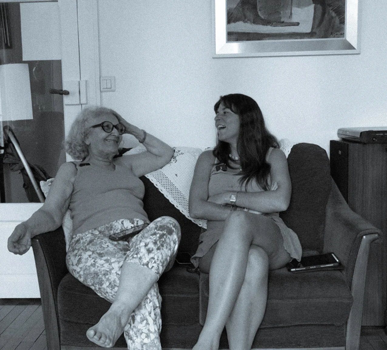 Deux femmes, une âgée et une plus jeune, assises sur un canapé souriant et riant ensemble dans un salon, en train de partager un moment de convivialité.