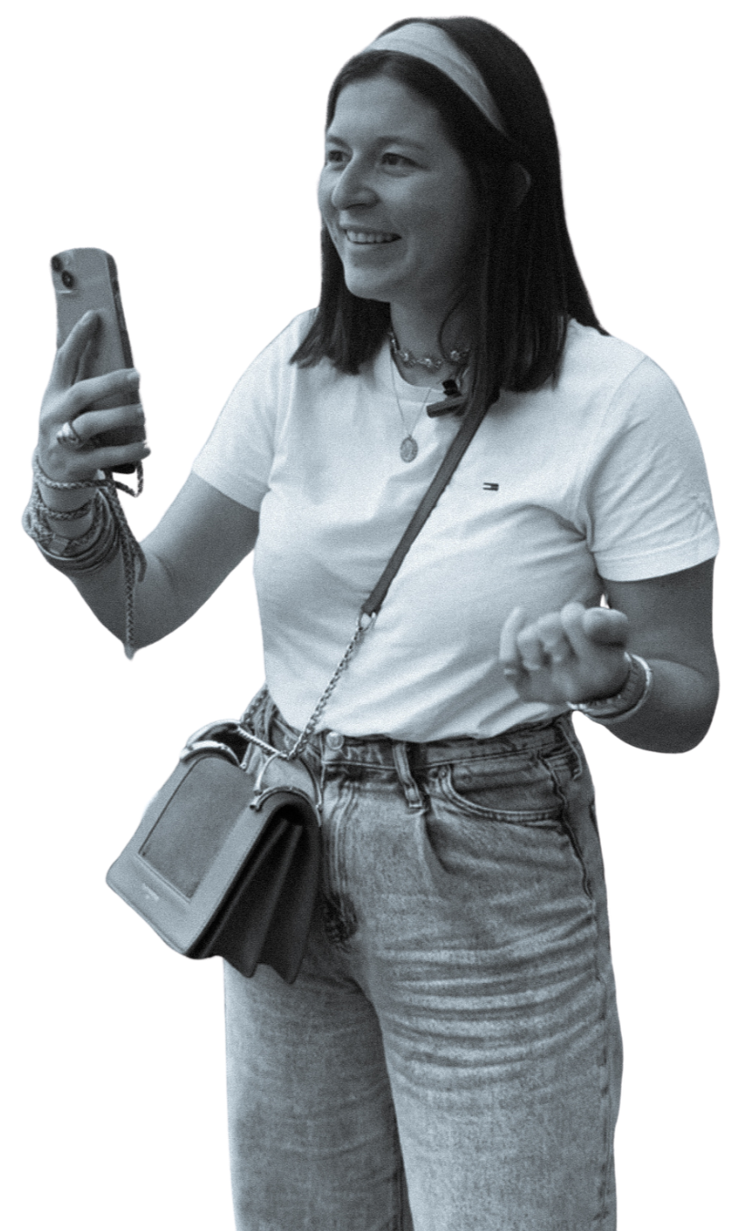 Jeune femme souriante prenant une vidéo avec son téléphone, elle interviewe quelqu'un, portant un T-shirt blanc, un jean, des bijoux et un sac à bandoulière.