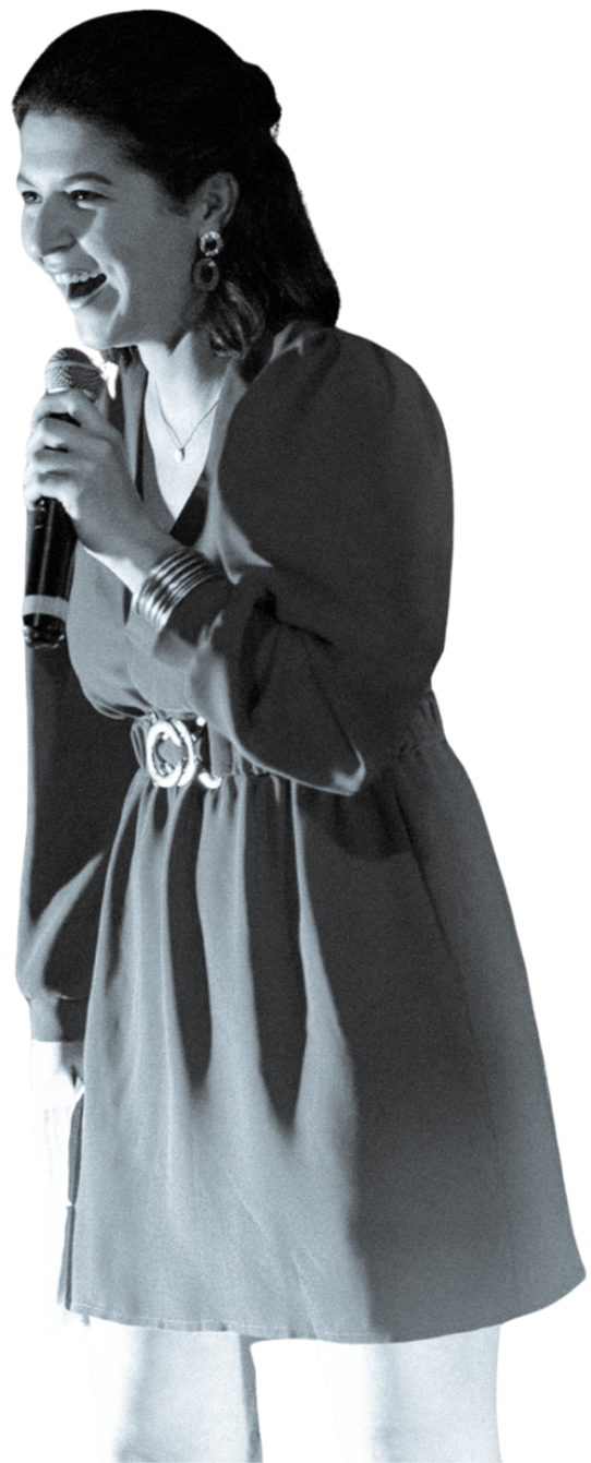 Femme souriante avec un micro, portant une robe et des bijoux, en train de parler ou de chanter, en noir et blanc, lors d'une performance, ou d'une présentation.