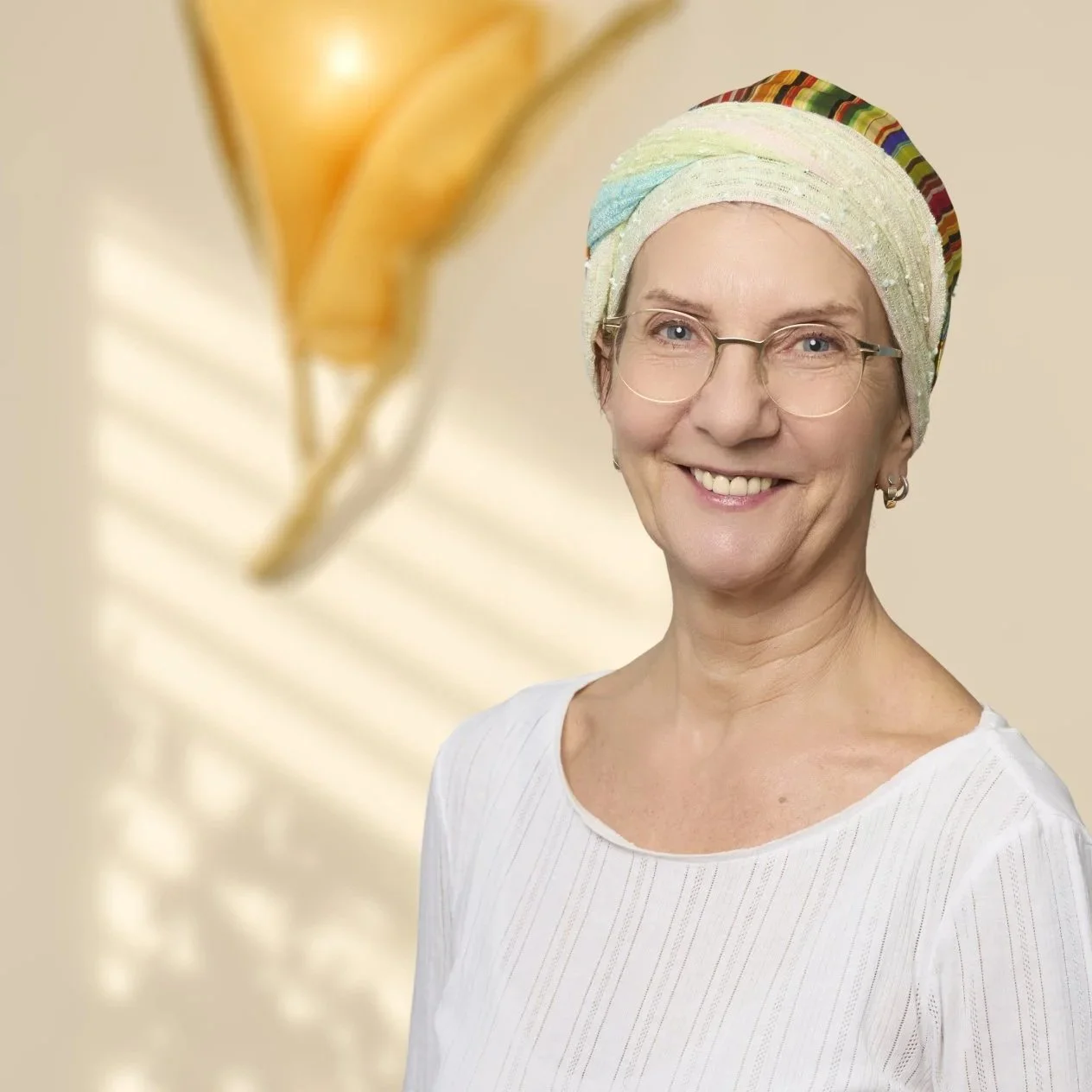 Eine lächelnde ältere Frau mit Brille, buntem Kopftuch und weißem Hemd, vor einem beigefarbenen Hintergrund mit einem goldfarbenen Balloon im Vordergrund.