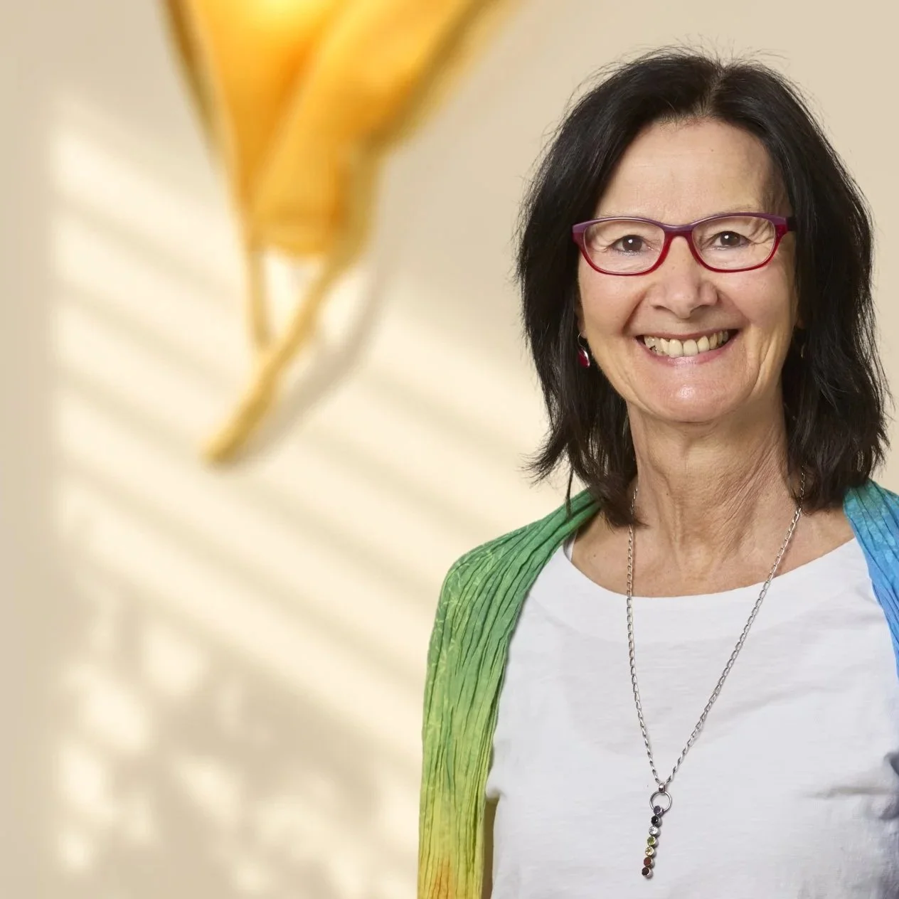 Eine lächelnde Frau mit dunklen Haaren, roten Brillen und buntem Schal vor einem neutralen Hintergrund.