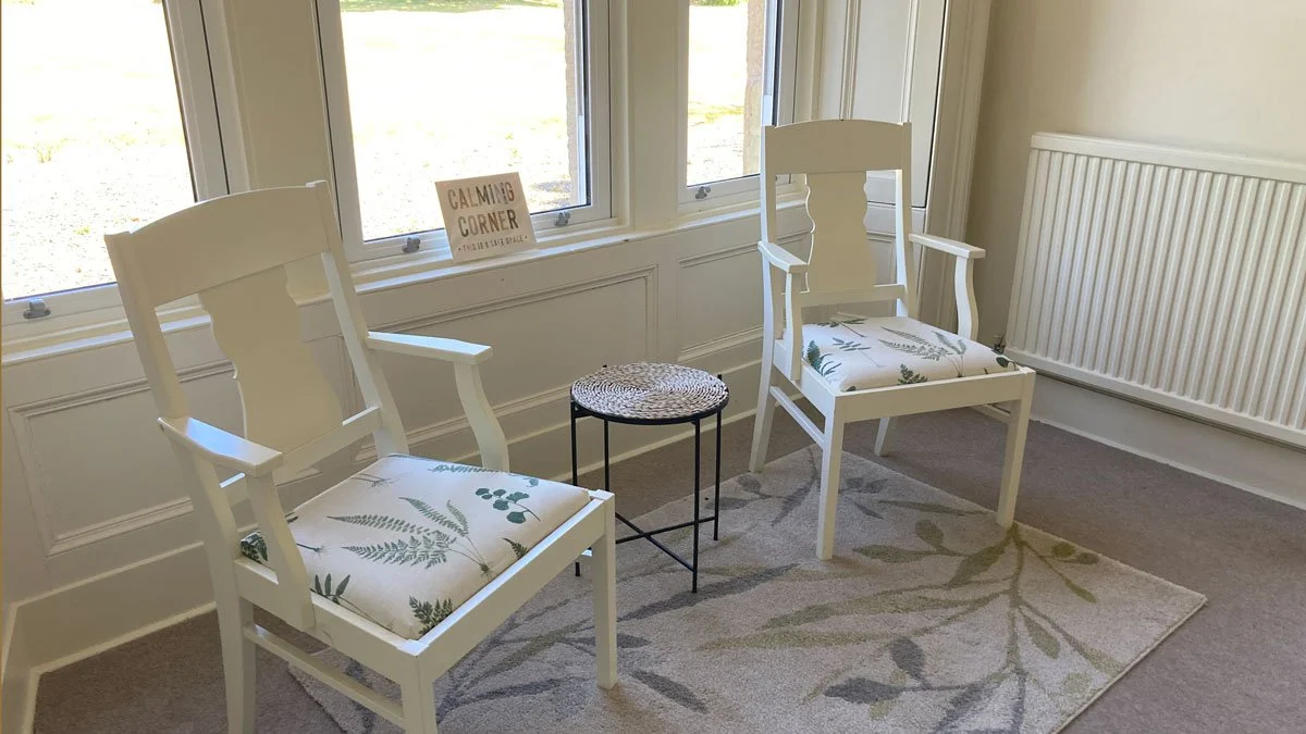 A cozy sitting area with two white wooden chairs with cushioned seats, a small round woven table in between, a beige rug with green leaf patterns, and a window with a sign that says "Calming Corner."