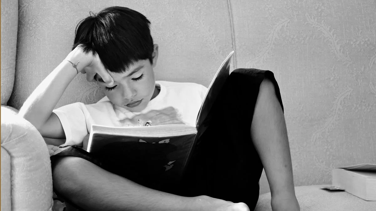A young boy with dark hair reading a book while sitting on a couch, resting his head on his hand.