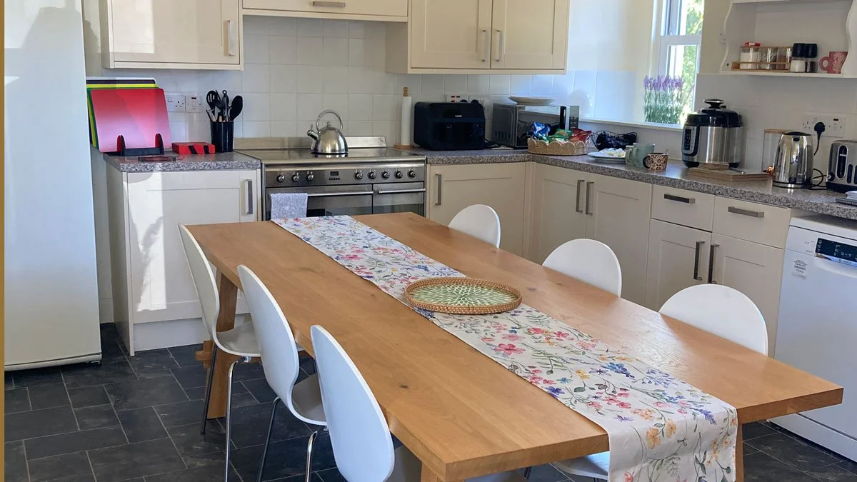Kitchen with a wooden dining table, six white chairs, a floral table runner, and various appliances on the counters including a toaster, kettle, and microwave.
