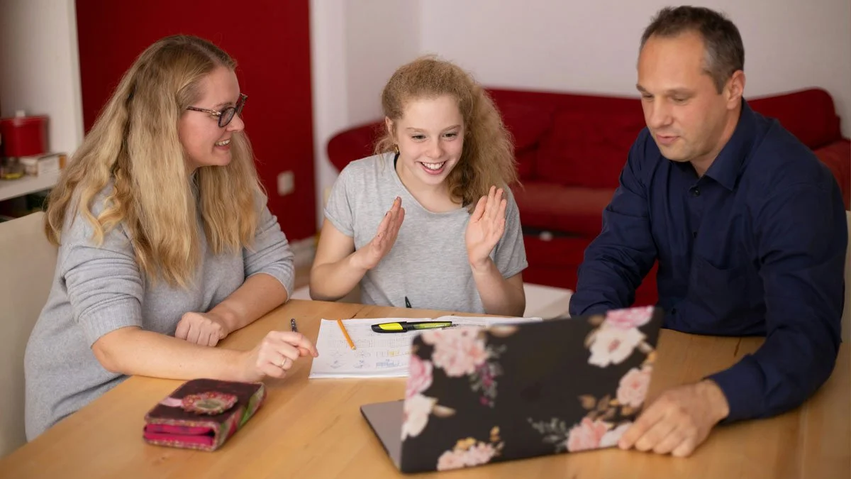 Family of three sitting at a table, looking at a laptop, smiling, with homework or papers in front of them in a cozy living room.