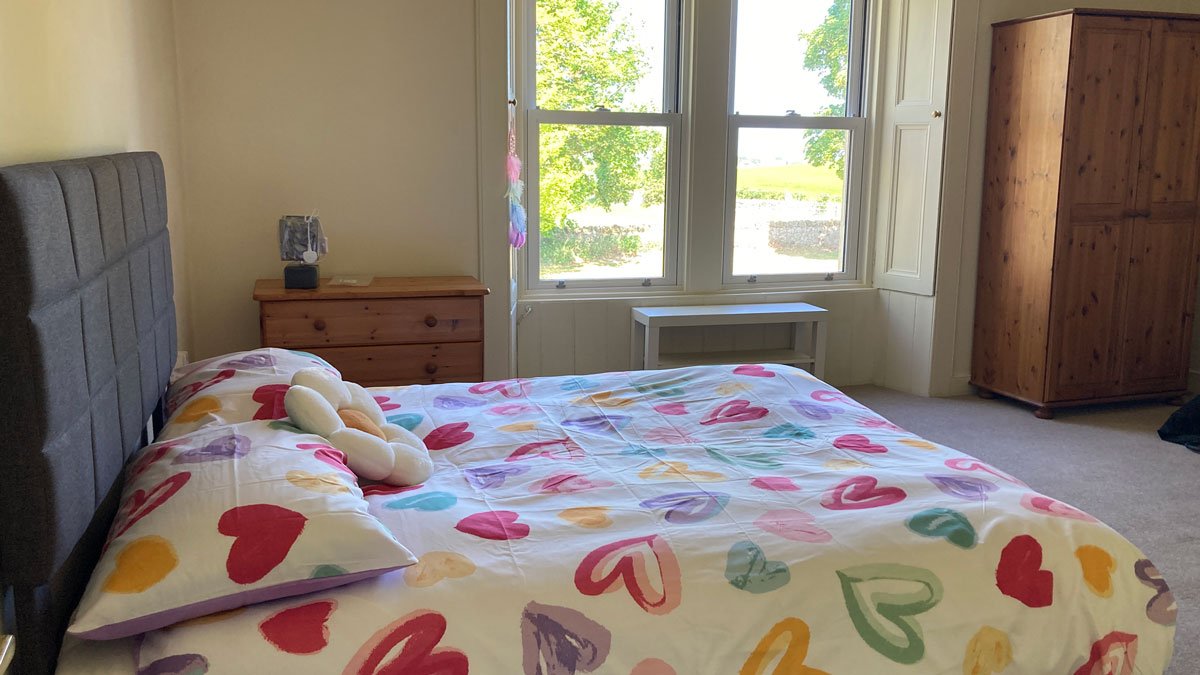 Bedroom with a bed covered in a colorful heart-patterned quilt, a gray headboard, a small pillow, a wooden nightstand with a lamp and some items, a large window with white trim and a view of greenery outside, a white bench below the window, and a wooden wardrobe.