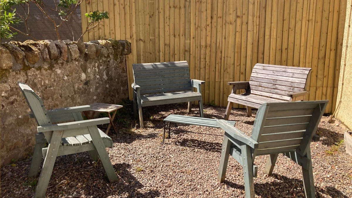 Outdoor patio with four wooden chairs, a small table, and a wooden fence, with a stone wall on the left side.