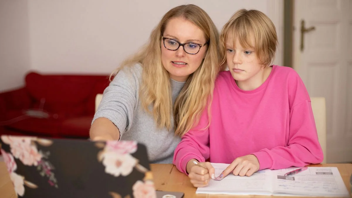 A woman and a young girl sit at a table looking at a laptop. The woman has blonde hair, glasses, and is wearing a gray shirt, while the girl has blonde hair and is wearing a pink sweatshirt. They appear to be studying or working on homework.
