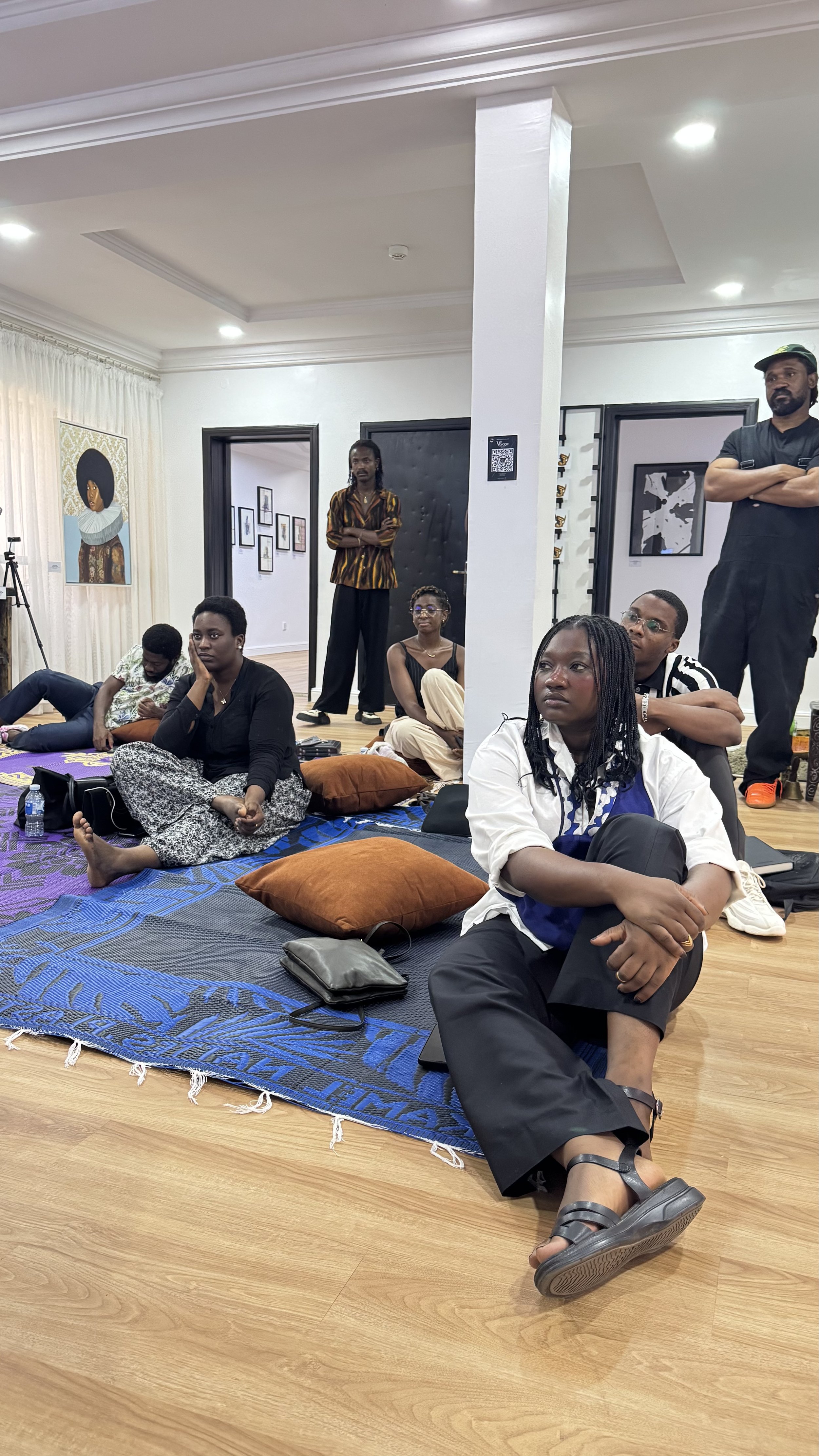 People sitting and standing inside an art gallery, with artworks and a tripod in the background.