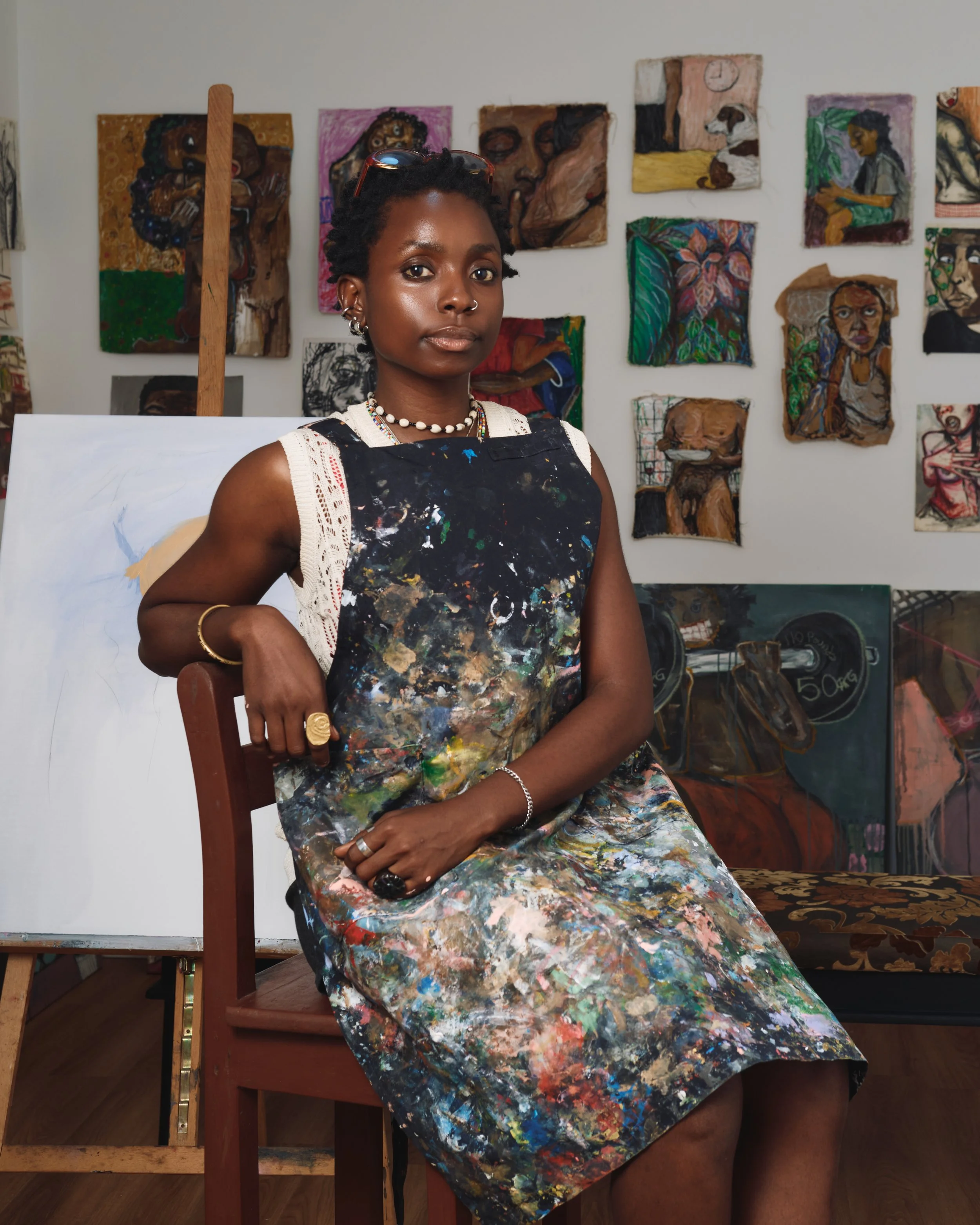 A young woman sitting on a chair in an art studio, wearing a paint-splattered apron, jewelry, and sunglasses on her head. She has short hair and is surrounded by colorful paintings on the walls and canvas behind her.