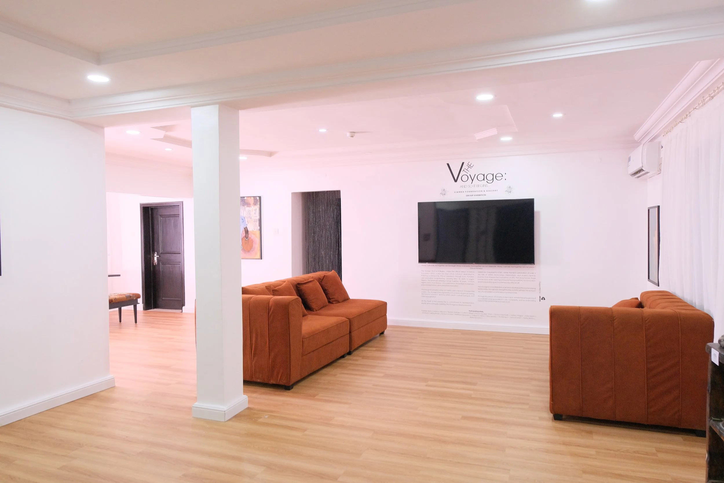 A living room with white walls, ceiling, and light wood flooring. Contains two orange sofas, a wall-mounted television, and artwork on the walls. There are recessed ceiling lights.
