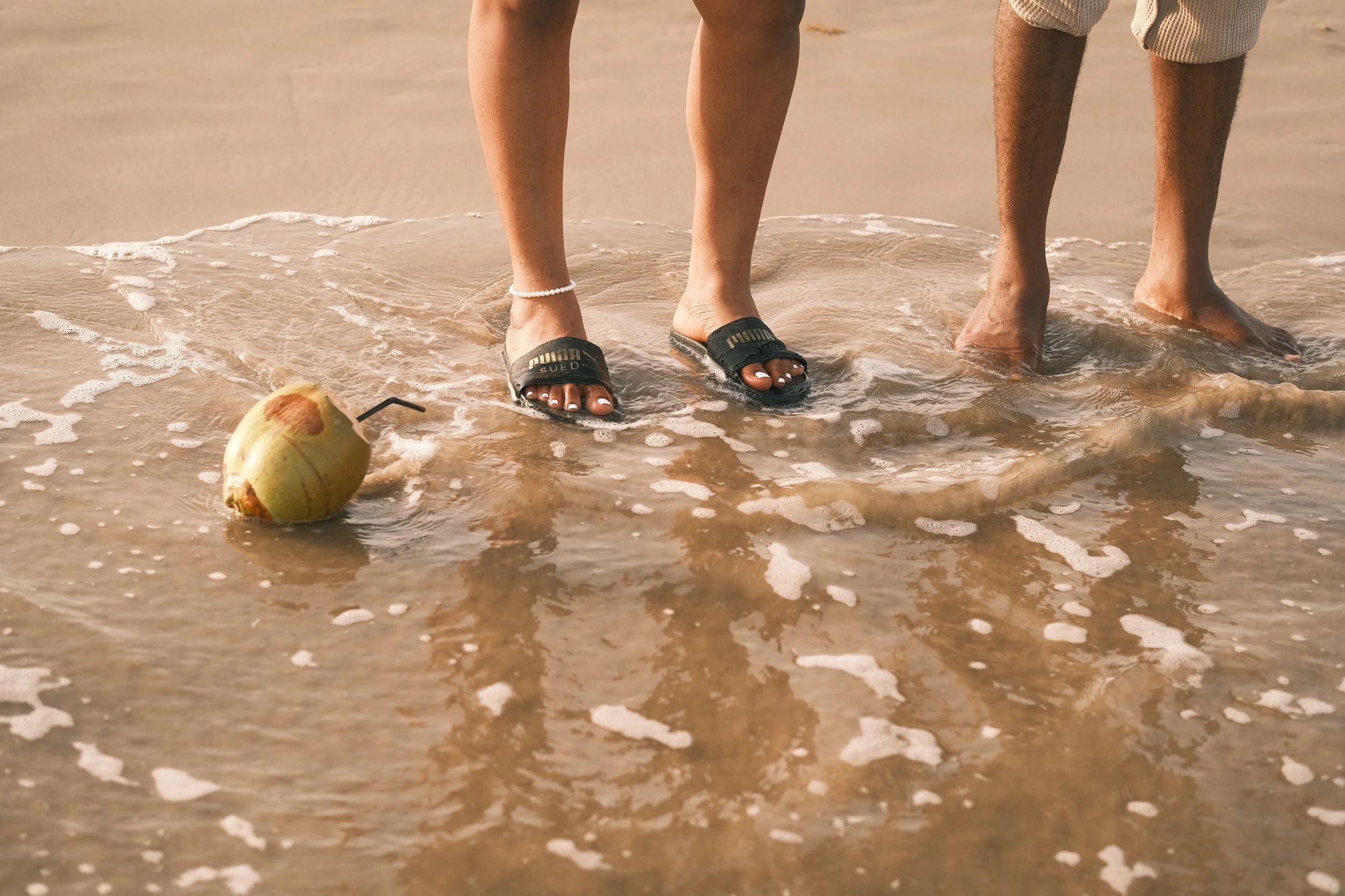 Two people standing in shallow water on a beach, with a coconut and straw in the water nearby.