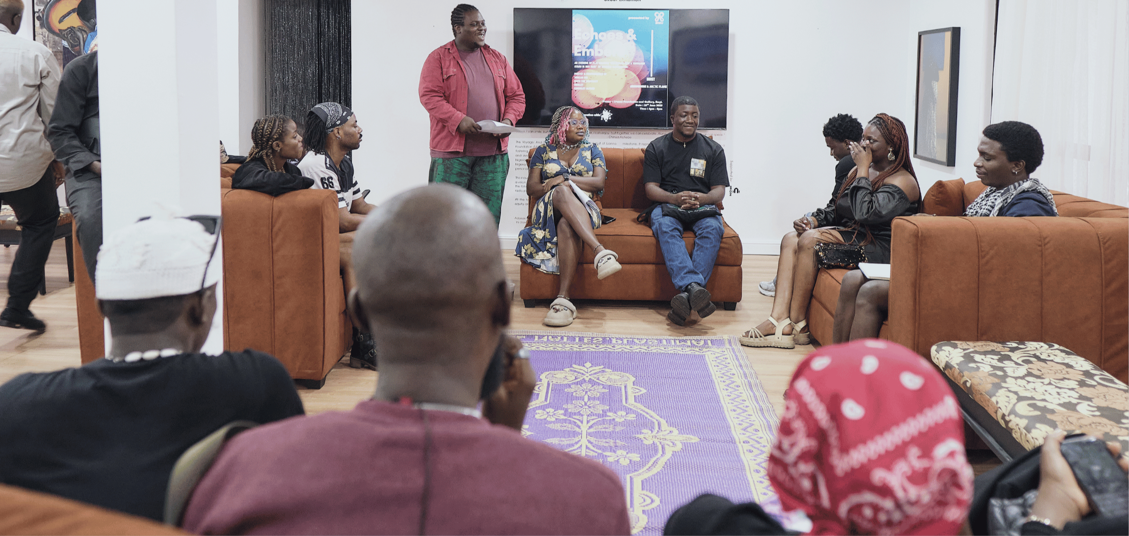 A group of people participating in a discussion or workshop in a room with comfortable seating, a purple rug, and a large screen displaying a presentation titled 'Energies & Embers' in the background. Some people are sitting on couches, listening attentively, while one woman stands holding papers.