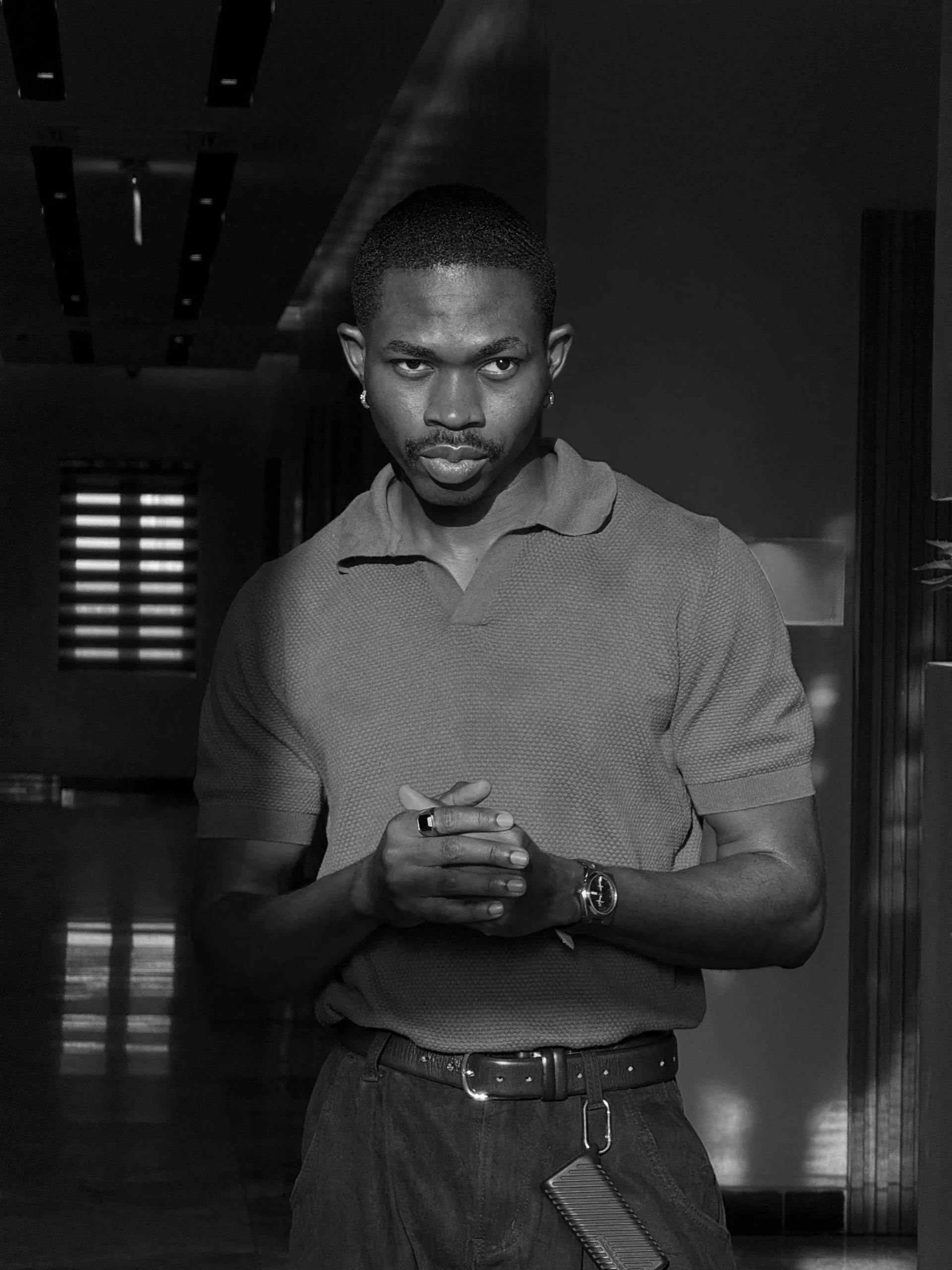 Black and white photo of a man with short hair, earrings, wearing a polo shirt, standing indoors with his hands clasped, looking at the camera.