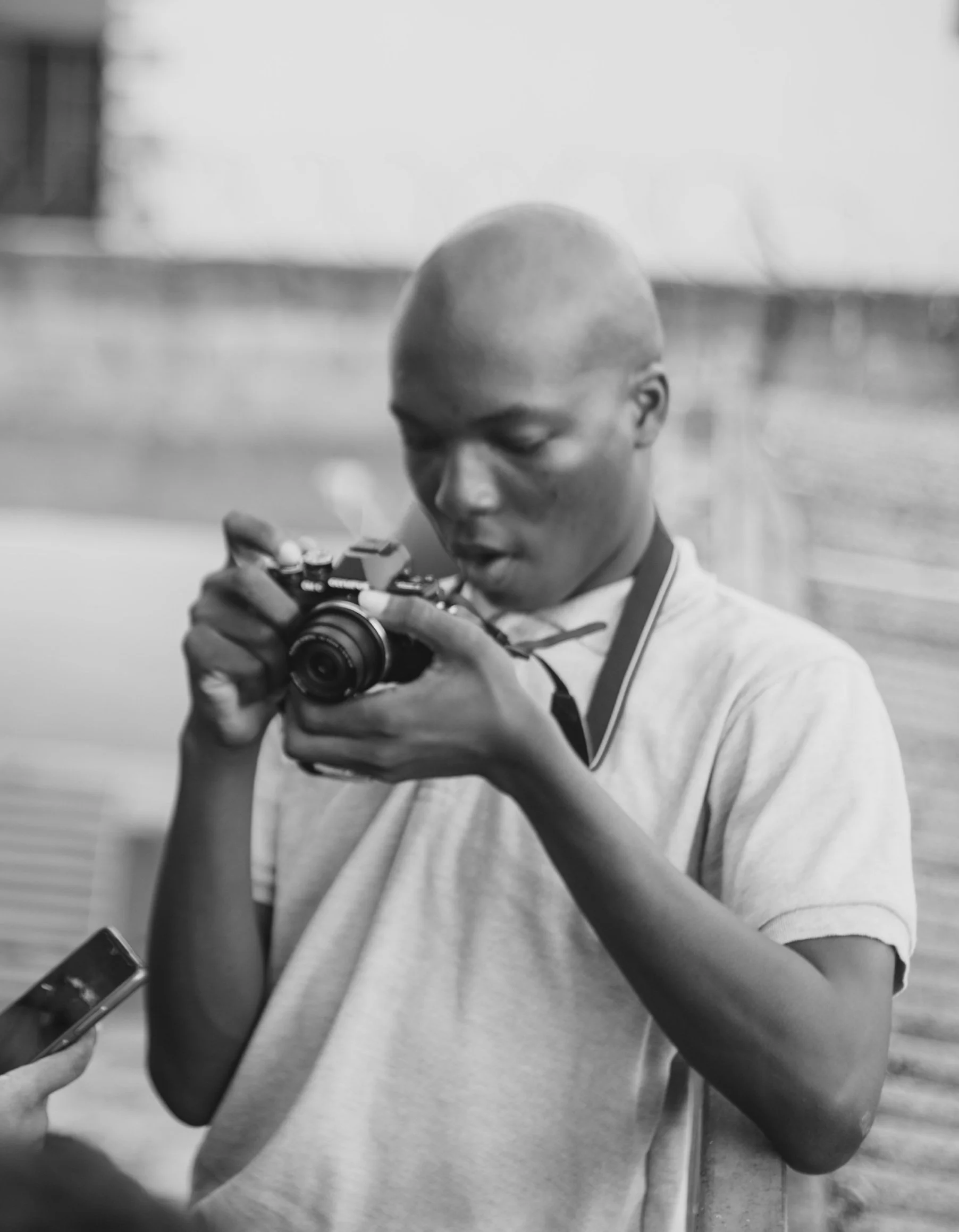 A young man with a shaved head looking at a camera and a person holding a phone