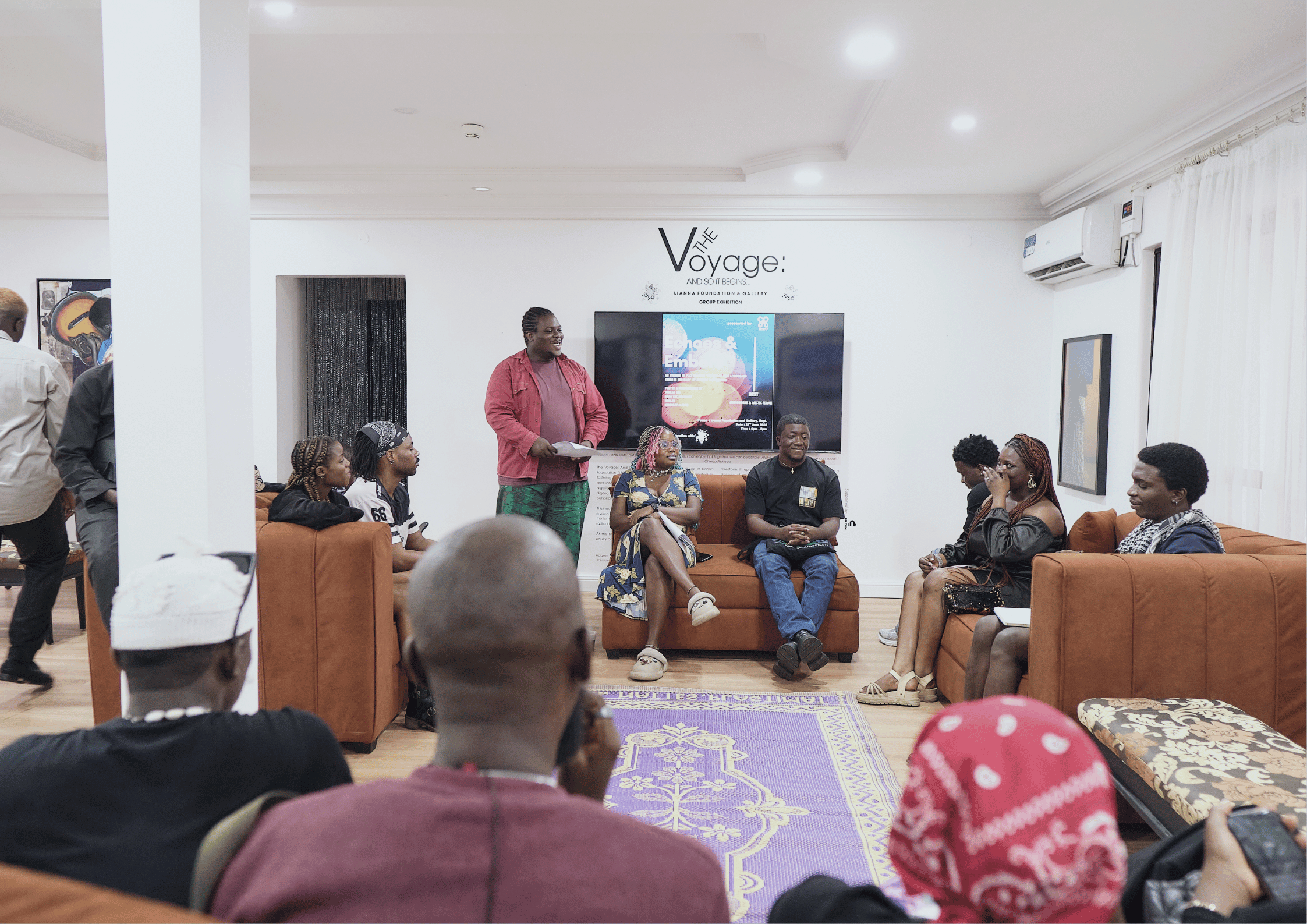 Group of people in a room attending a presentation or discussion, with some seated on sofas and others standing, in a well-lit room with artwork on the walls and a large screen displaying a presentation behind the speaker.
