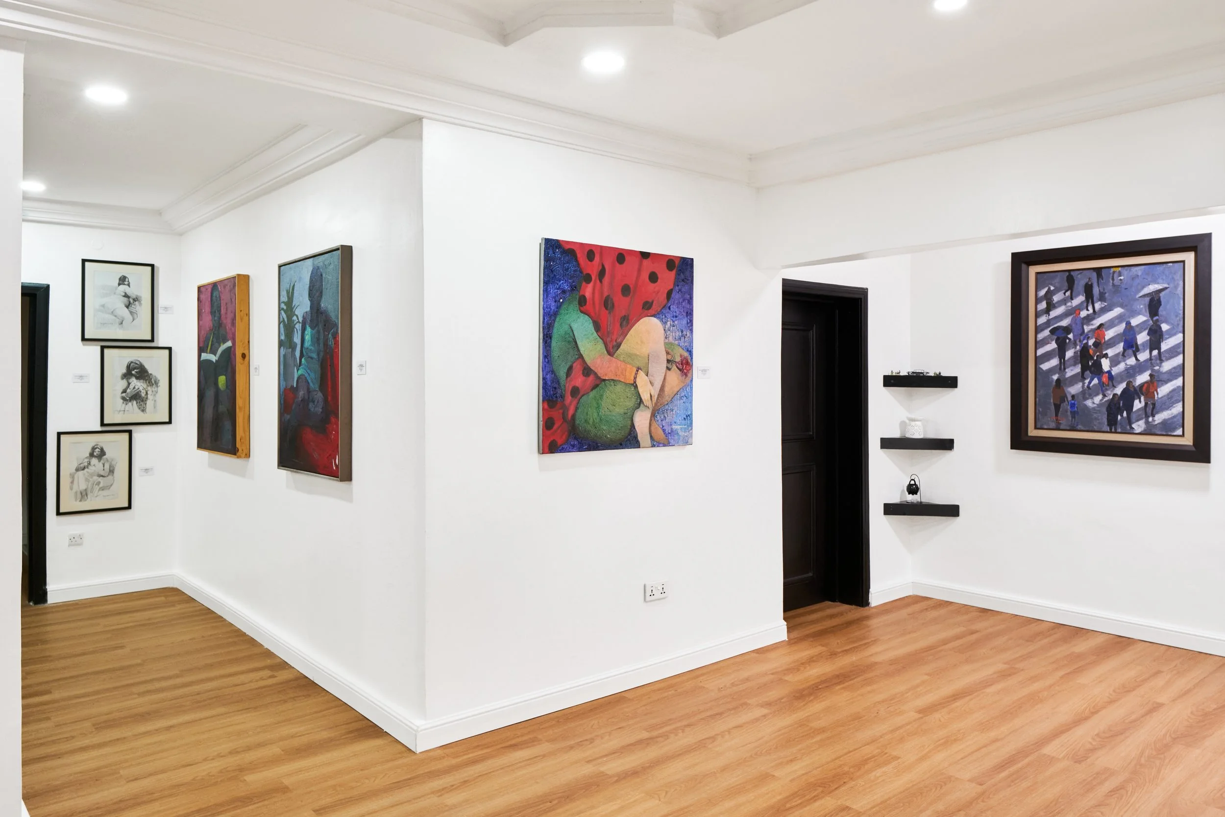 Interior view of an art gallery with white walls and wooden flooring, displaying various colorful paintings including portraits and abstract scenes.