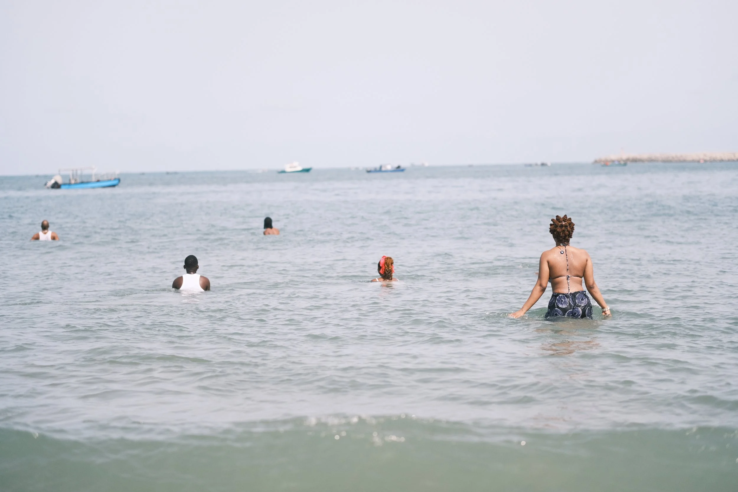People swimming and wading in the ocean during daytime, with boats visible in the background.