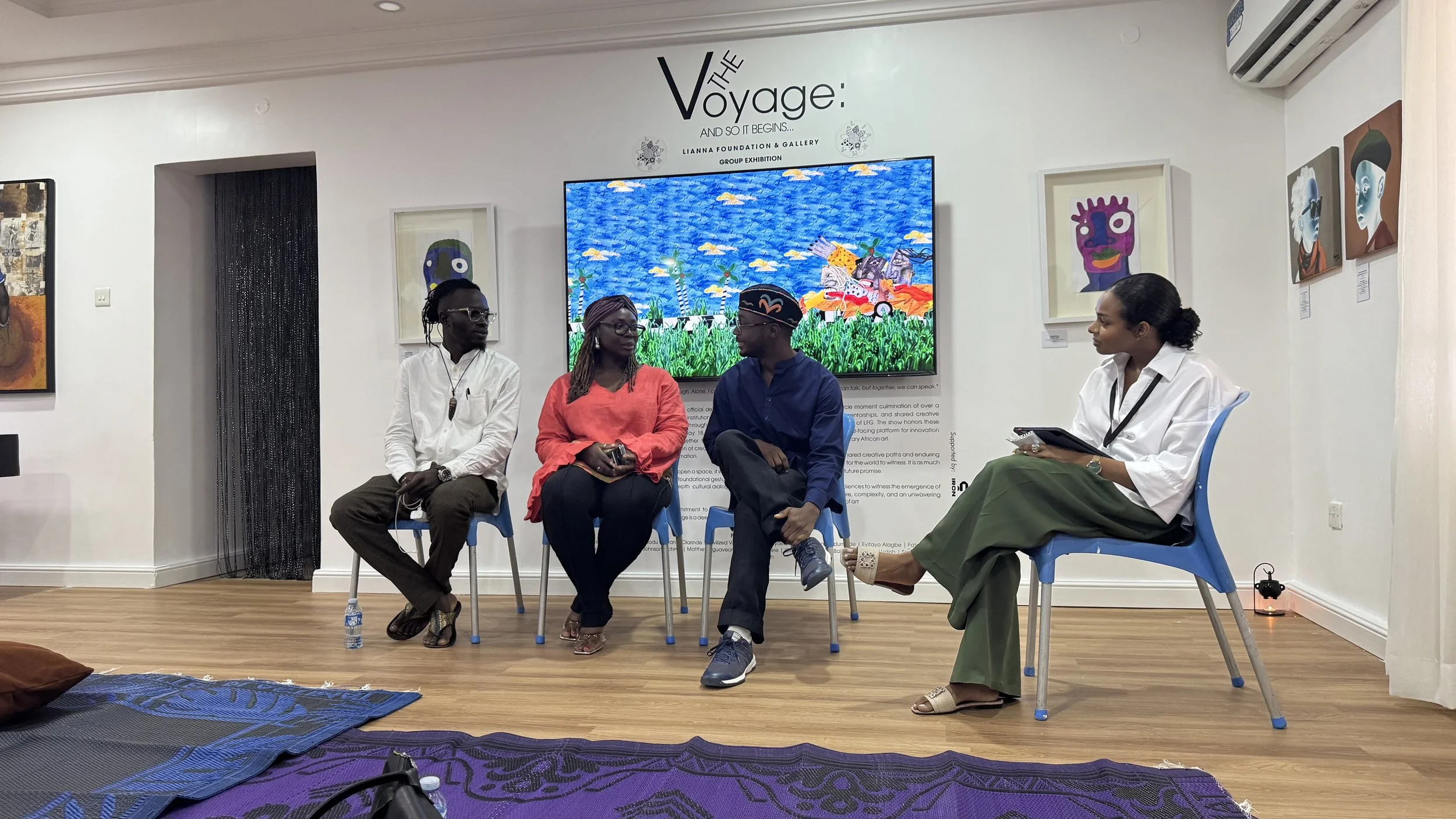 Four people participating in a panel discussion or interview in an art gallery, with colorful artwork displayed on the wall behind them.