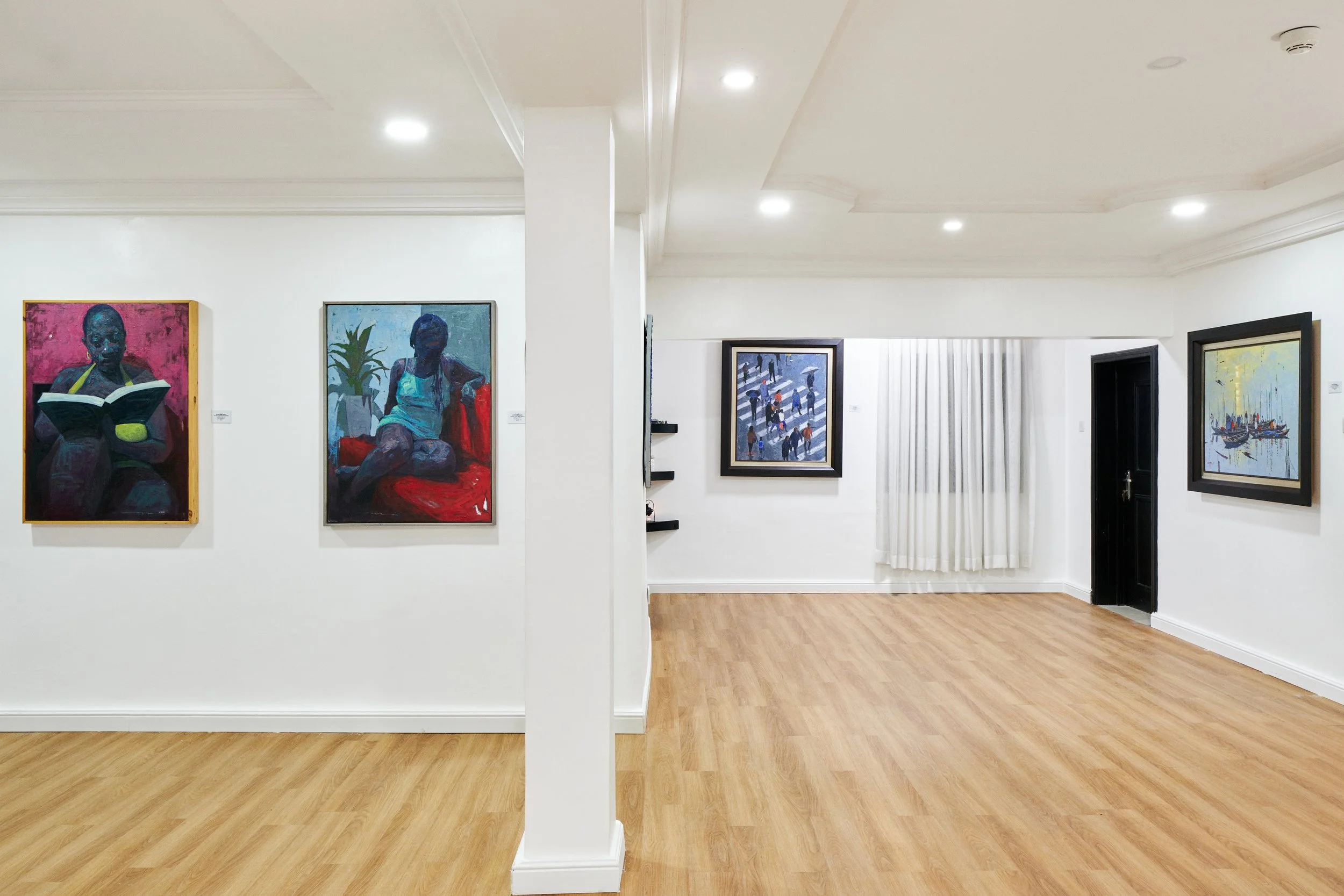 Interior view of an art gallery featuring paintings on white walls and a wooden floor, with a white curtain and black door at the back.