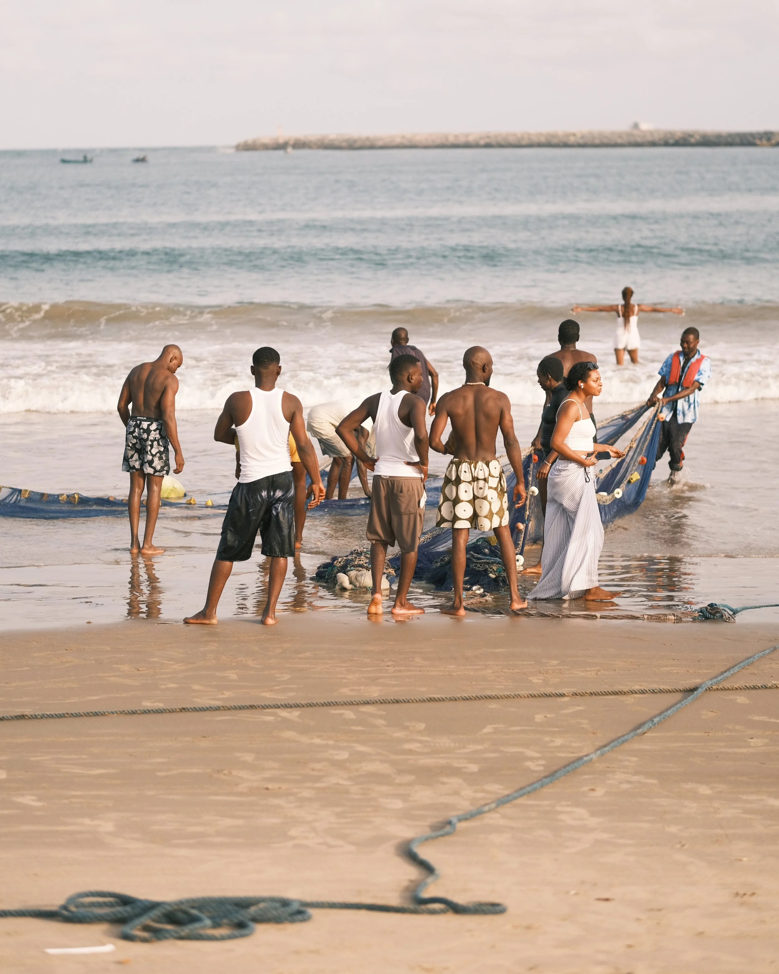 A group of people on a beach launching or retrieving a fishing net from the water with the ocean and a breakwater in the background.