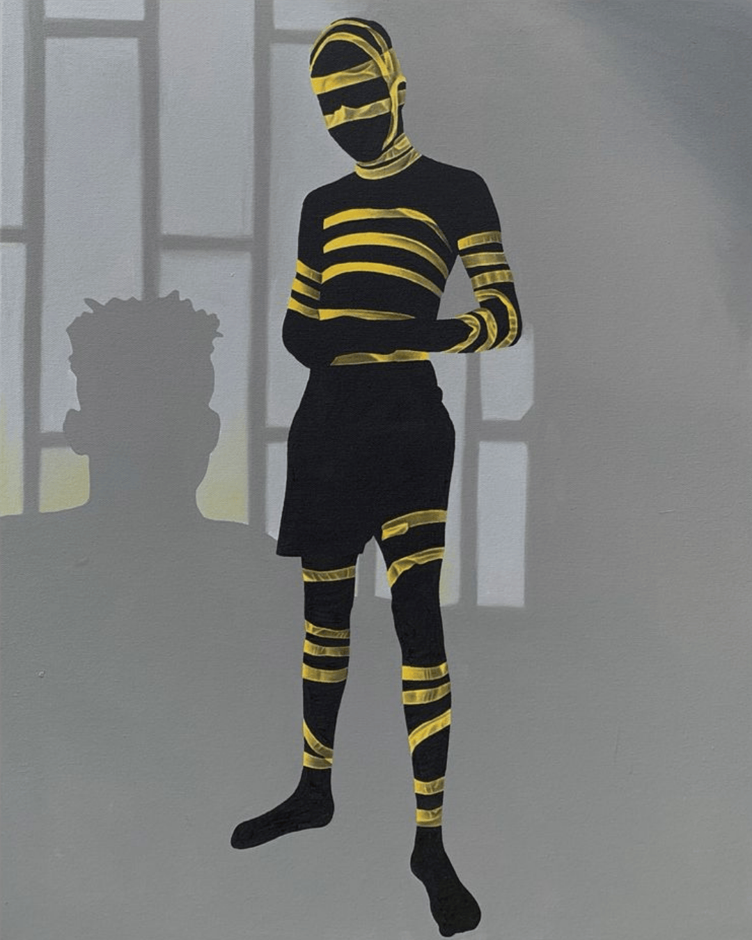 A person dressed in black with yellow striped tape wrapped around their body, face covered, standing with arms crossed indoor with windows in the background.