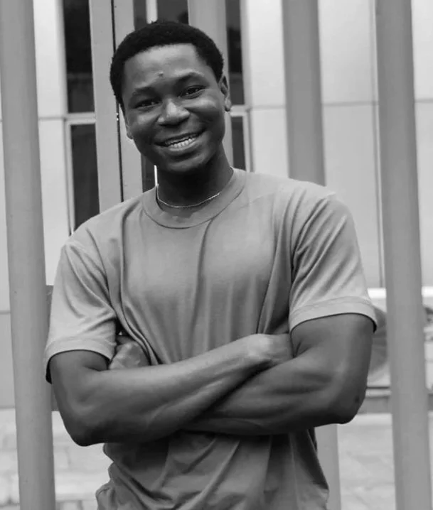 A young man smiling with arms crossed, standing outdoors in front of a modern building with vertical metal slats.