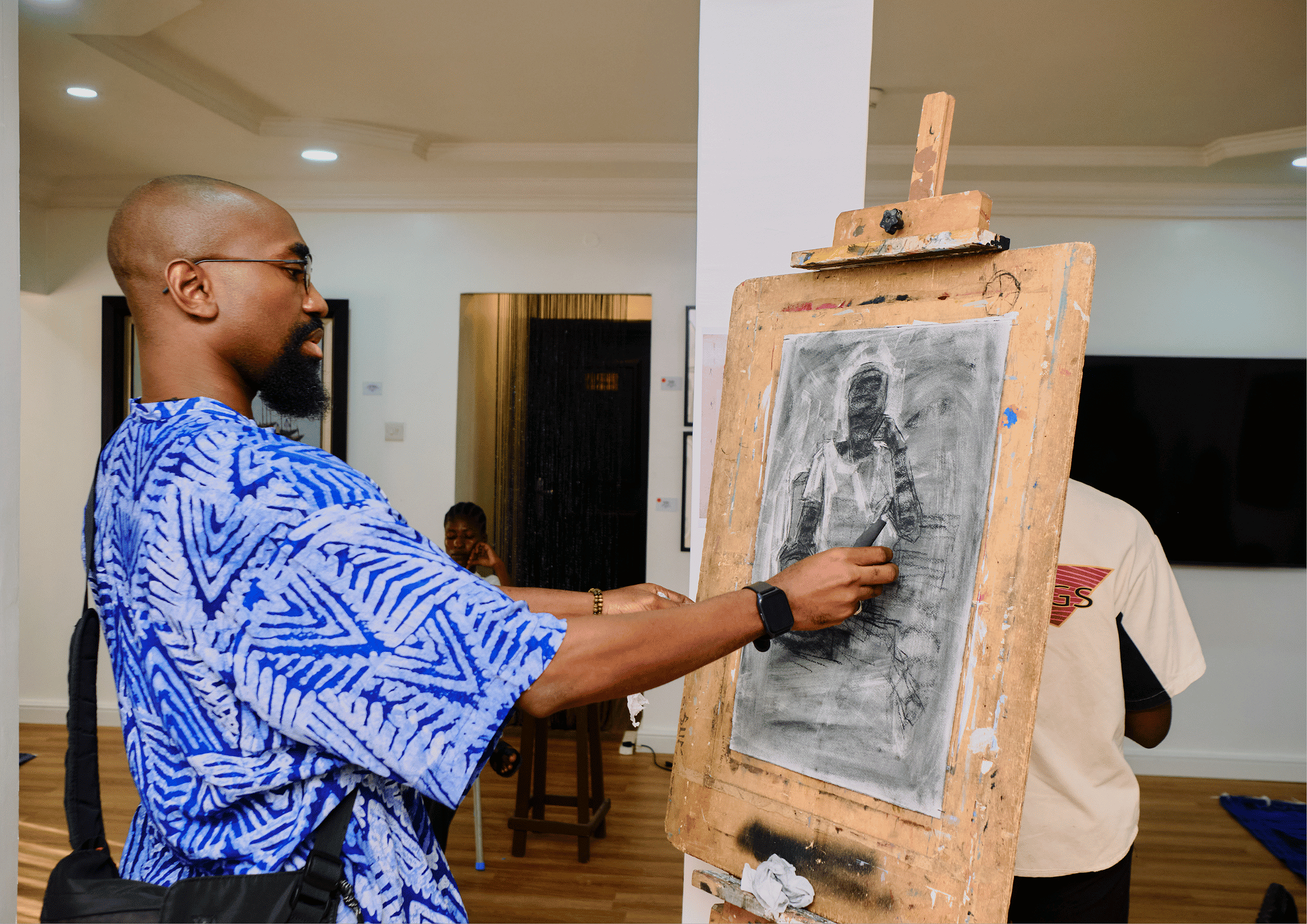 A man wearing glasses and a blue patterned shirt is drawing on a gray toned canvas with a charcoal pencil. The canvas is mounted on an easel in an indoor art gallery or studio. There is a woman in the background sitting at a table, and the room has white walls and wooden flooring.