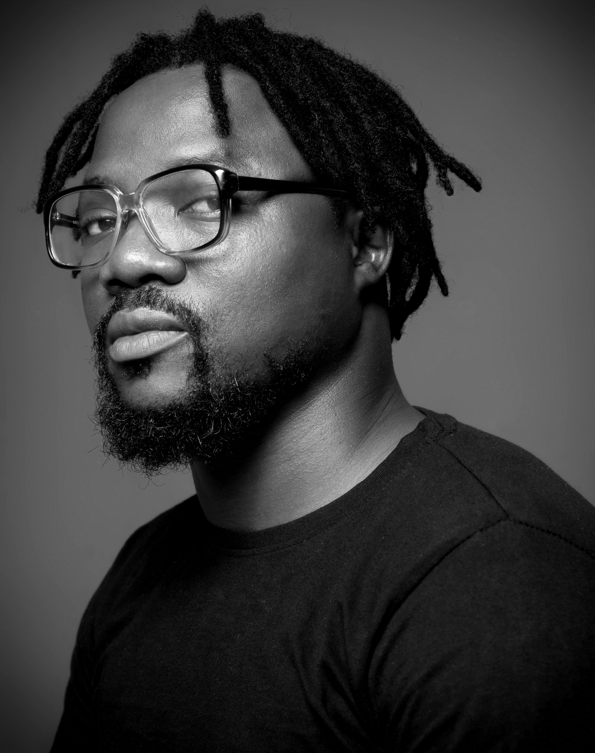 Black and white portrait of an African American man with dreadlocks, beard, wearing glasses and a black shirt, looking at the camera.