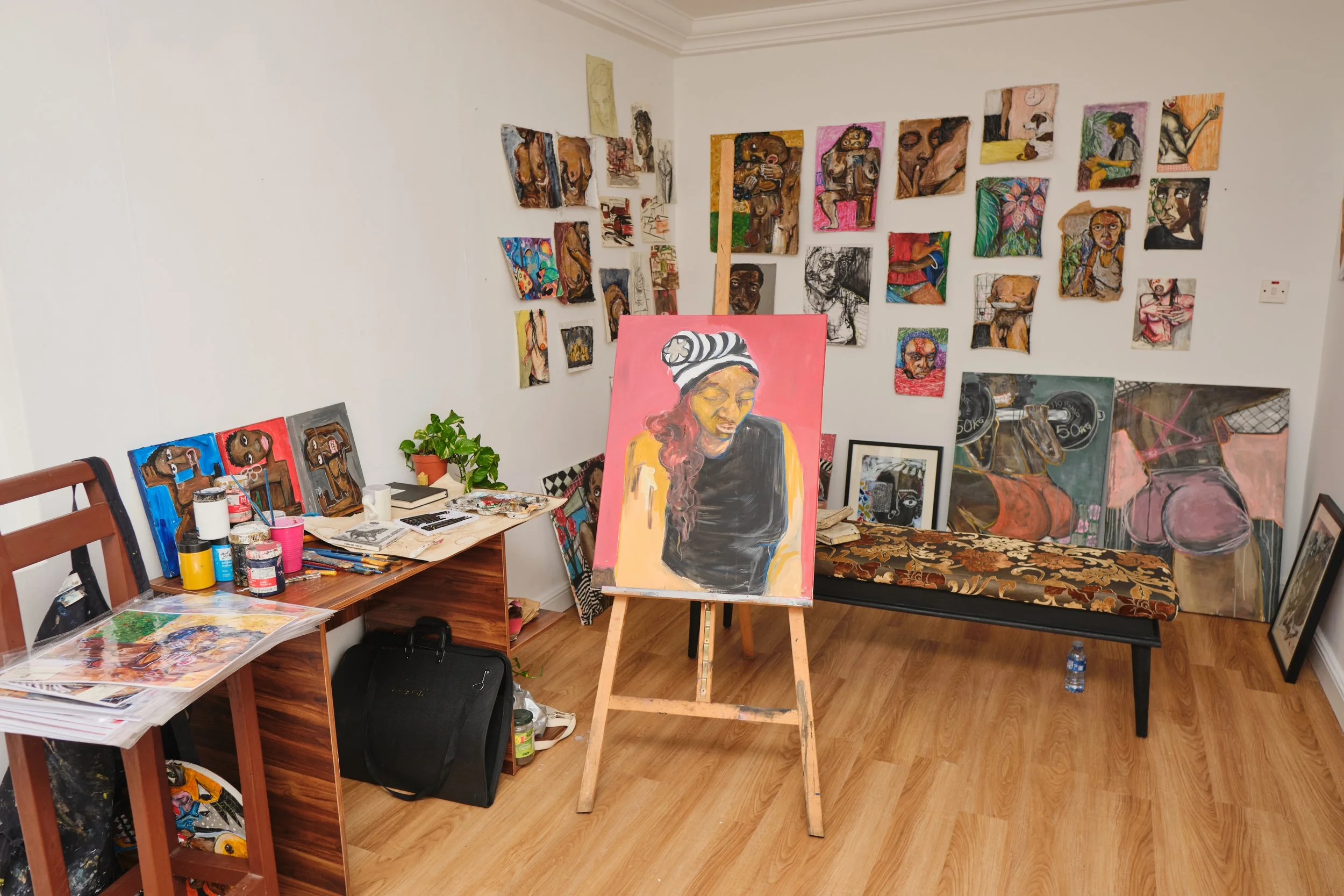 Artist's studio with various abstract and portrait paintings on walls and easel, artwork on tables, and art supplies, with a portrait of a woman in the center.