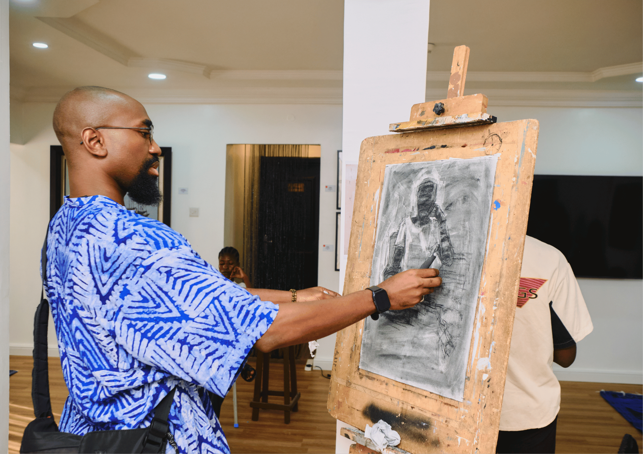 A man with a beard and glasses, wearing a blue patterned shirt, is sketching on a grayscale drawing on an easel in an art gallery, with people in the background.