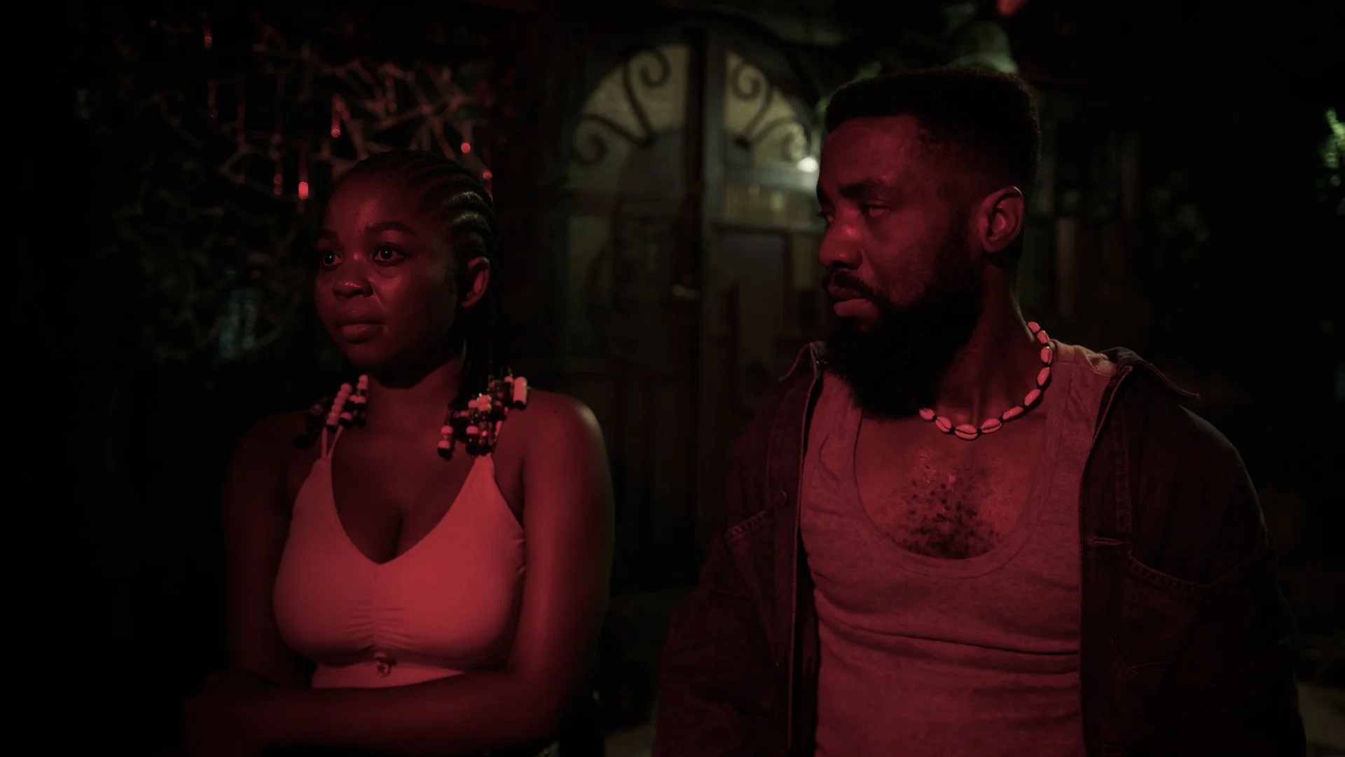 A young woman and a man sit side by side in a dimly lit setting with a dark background. The woman has braided hair with beads and is wearing a sleeveless top, while the man has a beard, short hair, and wears a shell necklace and a vest over a t-shirt. Both are looking to their left with serious expressions, illuminated by red lighting.