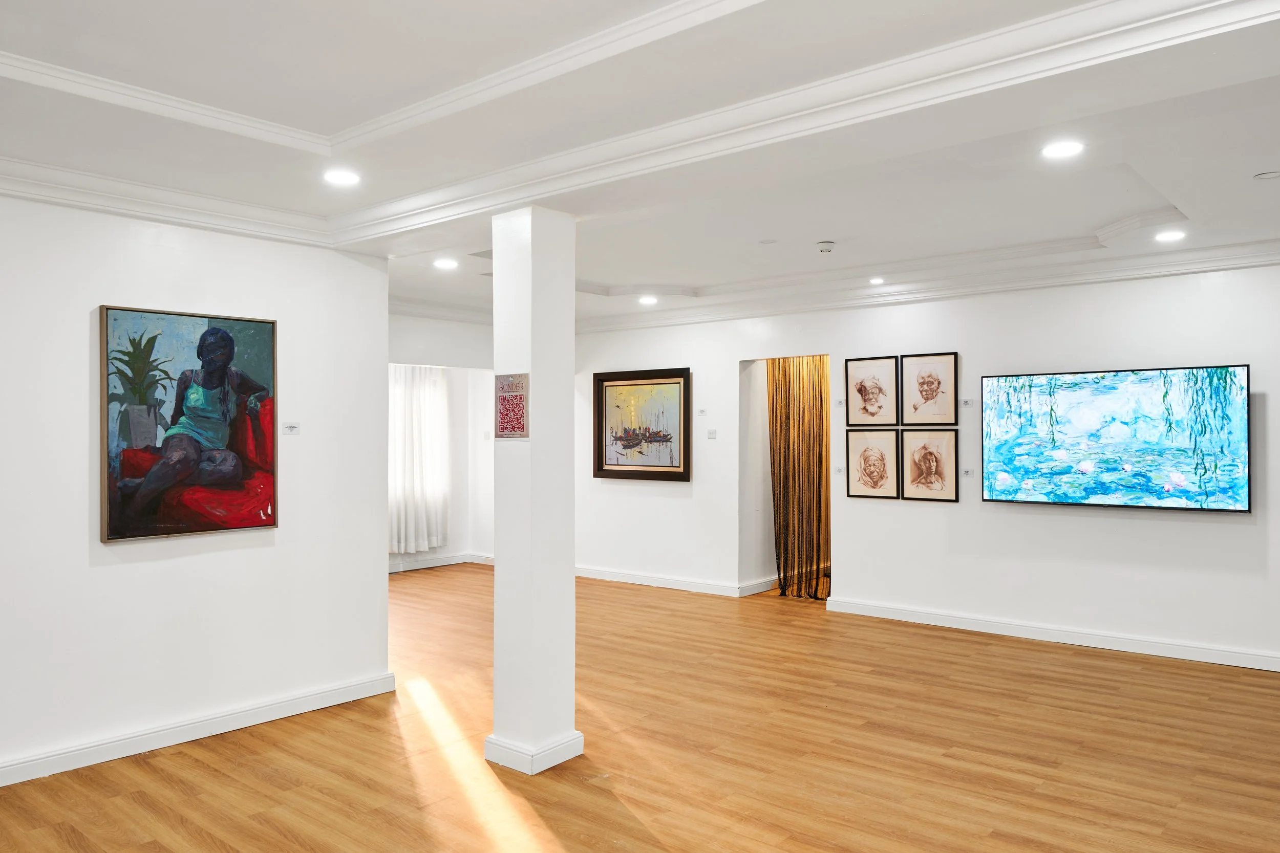 Art gallery with white walls, wooden floors, and multiple paintings, including a portrait of a woman on the left, a framed landscape in the center, a set of four sketches on the right, and a digital screen displaying an abstract water scene