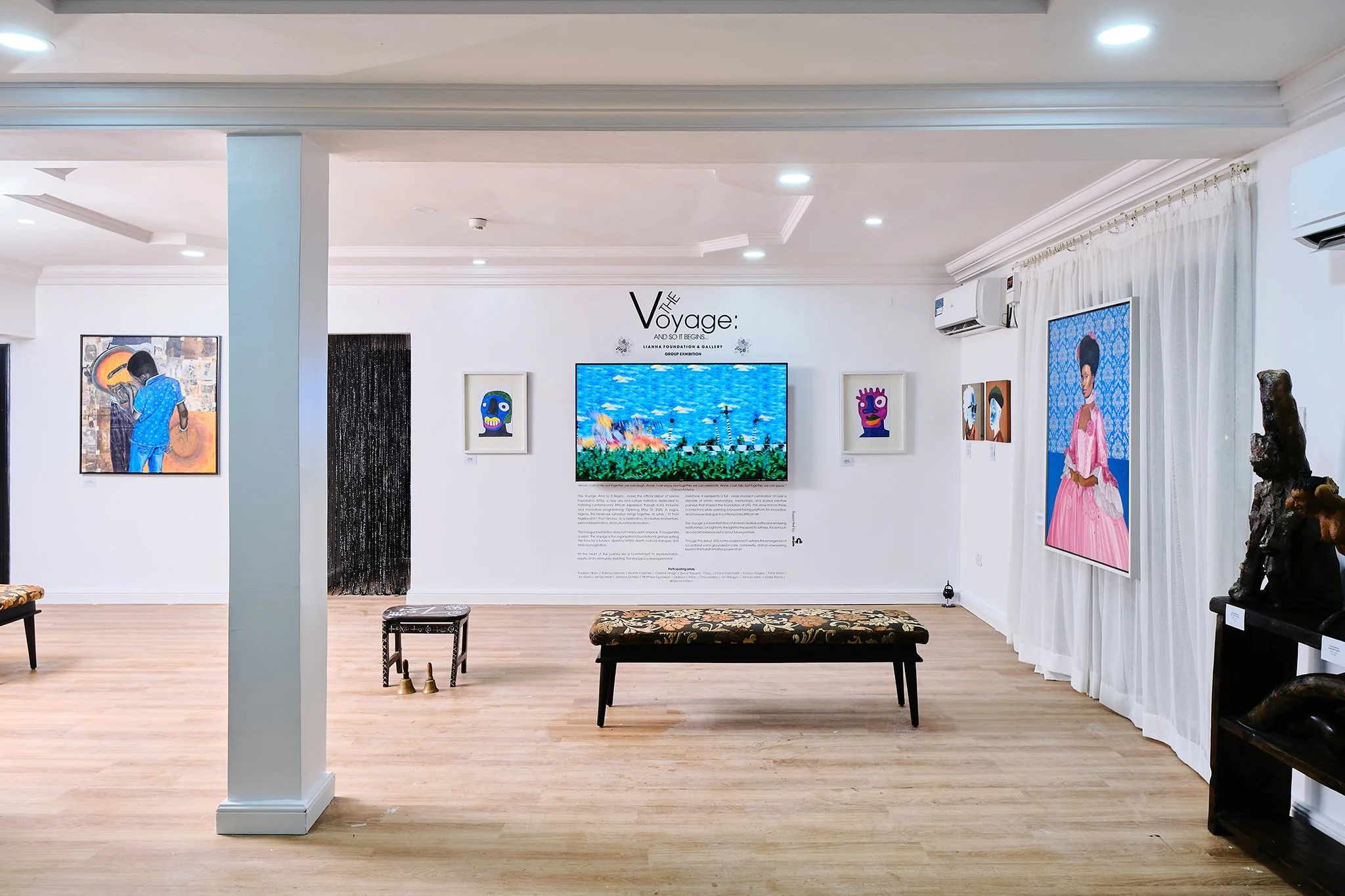 Art gallery with paintings and digital art displayed on white walls, benches in the center, and wooden flooring.