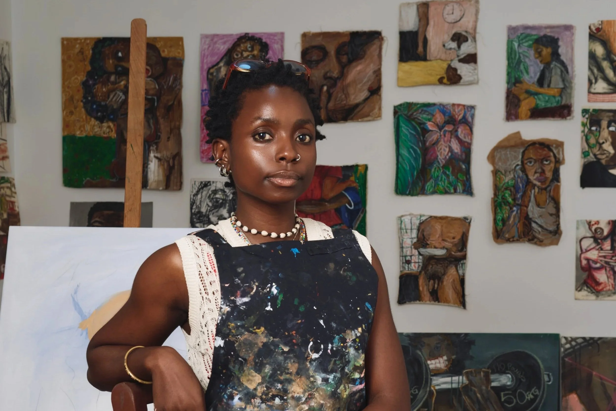 A young woman with short black hair decorated with sunglasses on her head, wearing a beaded necklace and earrings, standing in front of a wall of colorful paintings in an art studio.