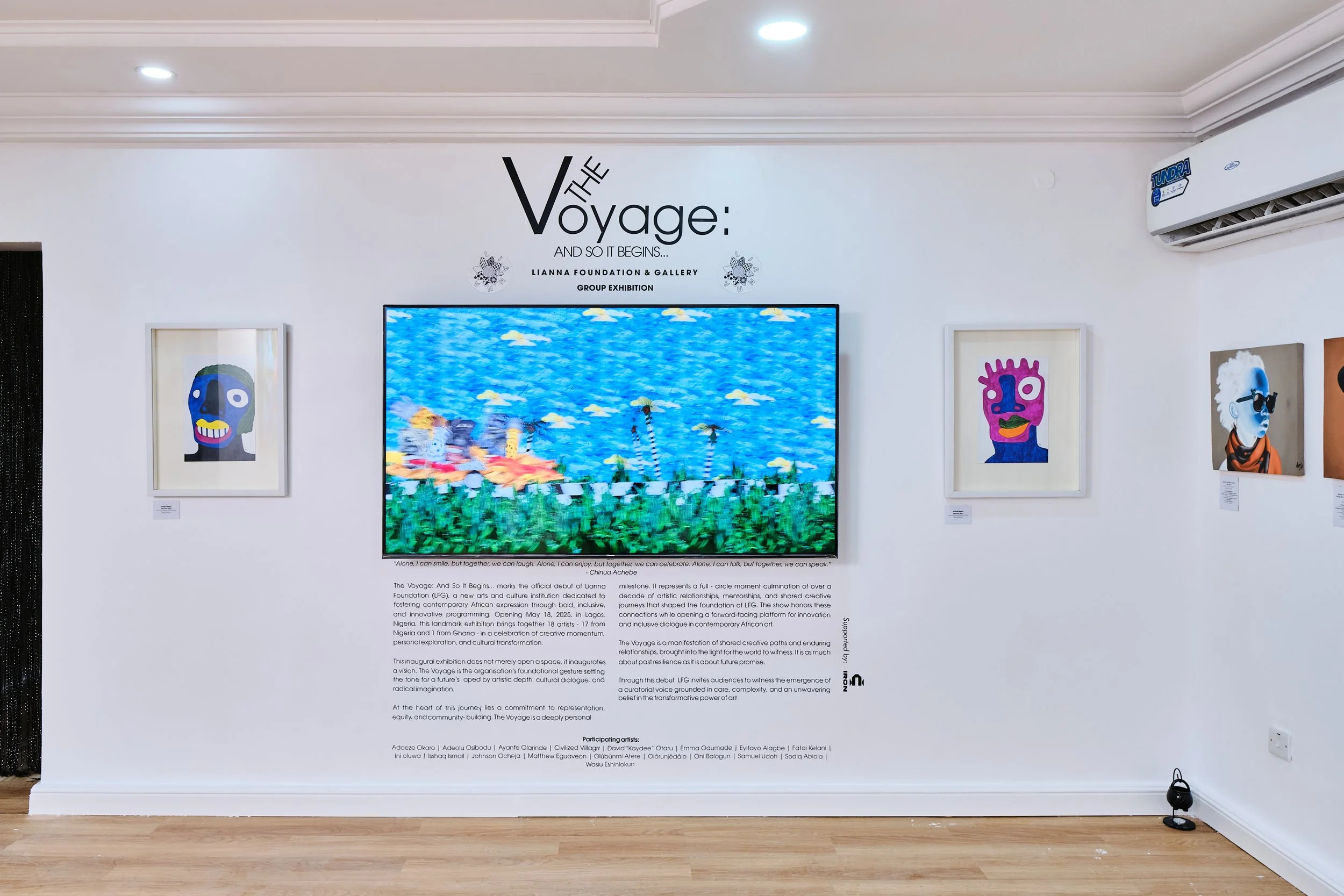 Art exhibition titled 'The Voyage: And So It Begins...' with a central digital display and framed artworks on white walls. The digital display shows a colorful, abstract landscape with blue sky and green grass. Framed artworks to the left and right feature stylized, colorful portraits with bold features.