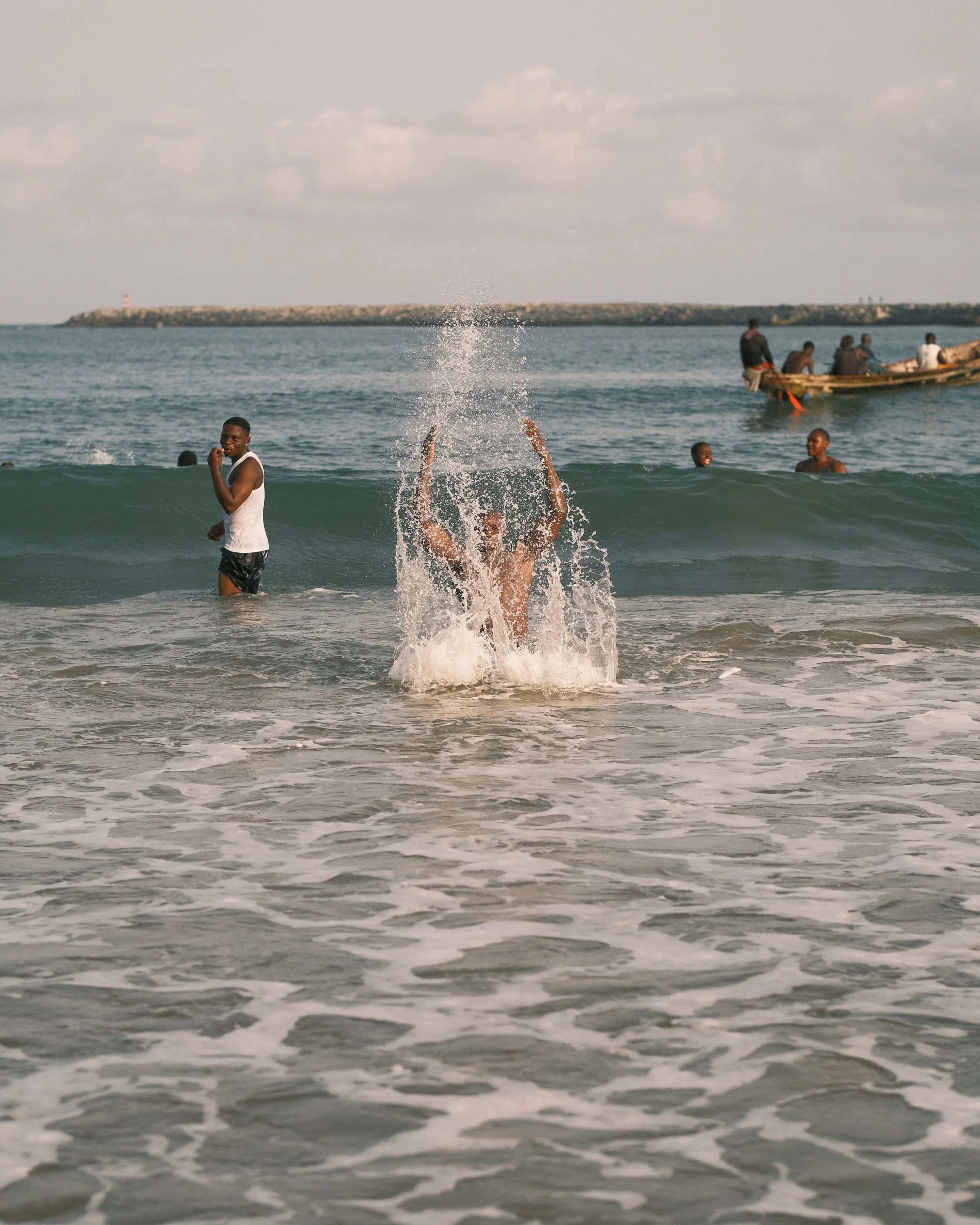 People enjoying time at the beach, some standing in the water, one person splashing water, a boat with passengers in the background.