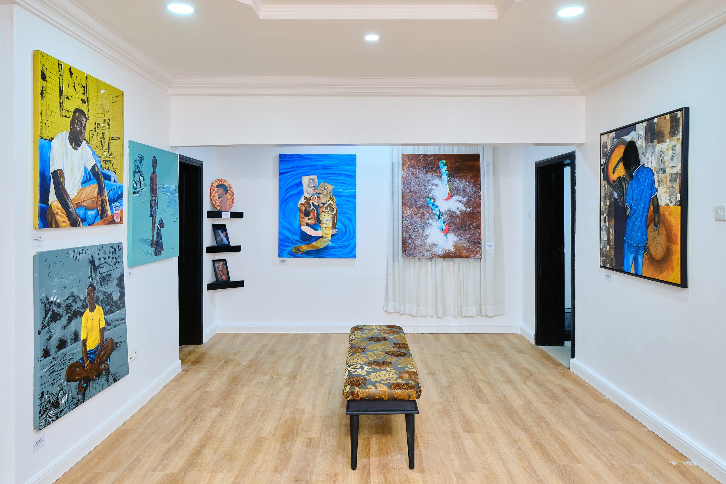 An art gallery room with several colorful paintings on white walls, a floral-patterned bench in the center, and light wooden flooring. The paintings depict various subjects, including people and abstract scenes.