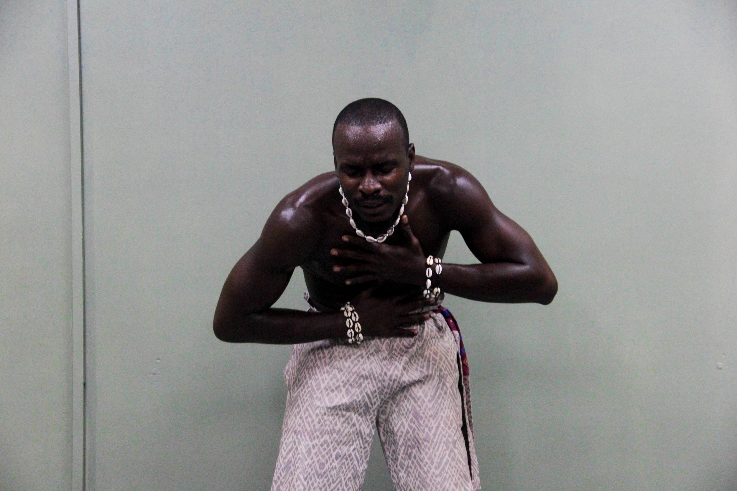 A shirtless man wearing traditional attire, including patterned pants and shell jewelry, bowing slightly with one hand on his chest and the other on his abdomen against a plain light grey background.