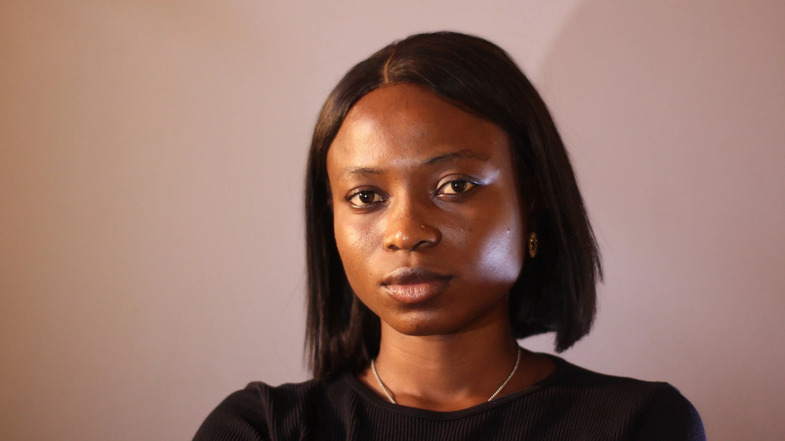 A young woman with medium-length black hair, wearing a black top, earrings, and a silver necklace, looking directly at the camera with a neutral expression.