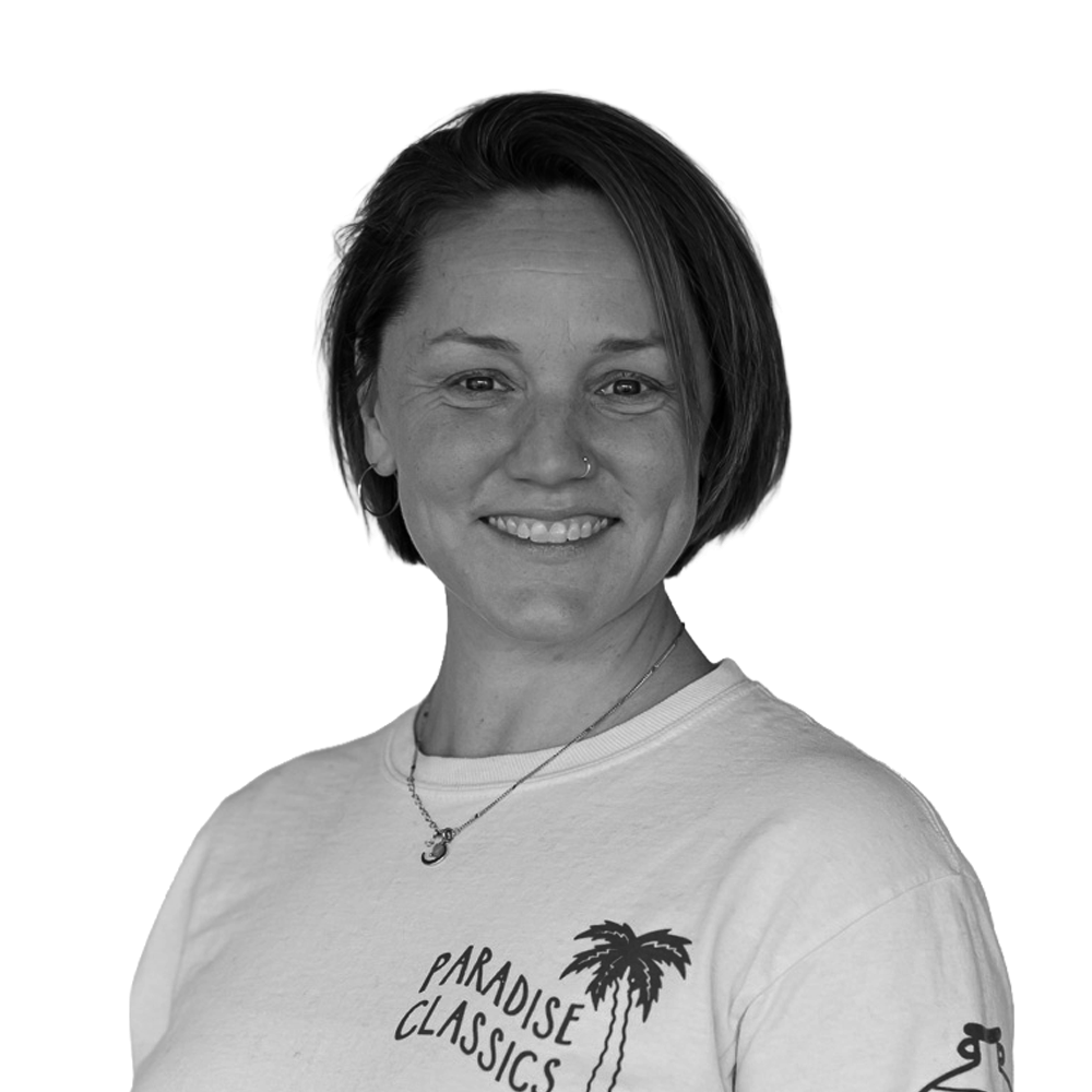 A woman with short dark hair, smiling, wearing a T-shirt with a palm tree graphic and the words 'Paradise Classics,' and a necklace.
