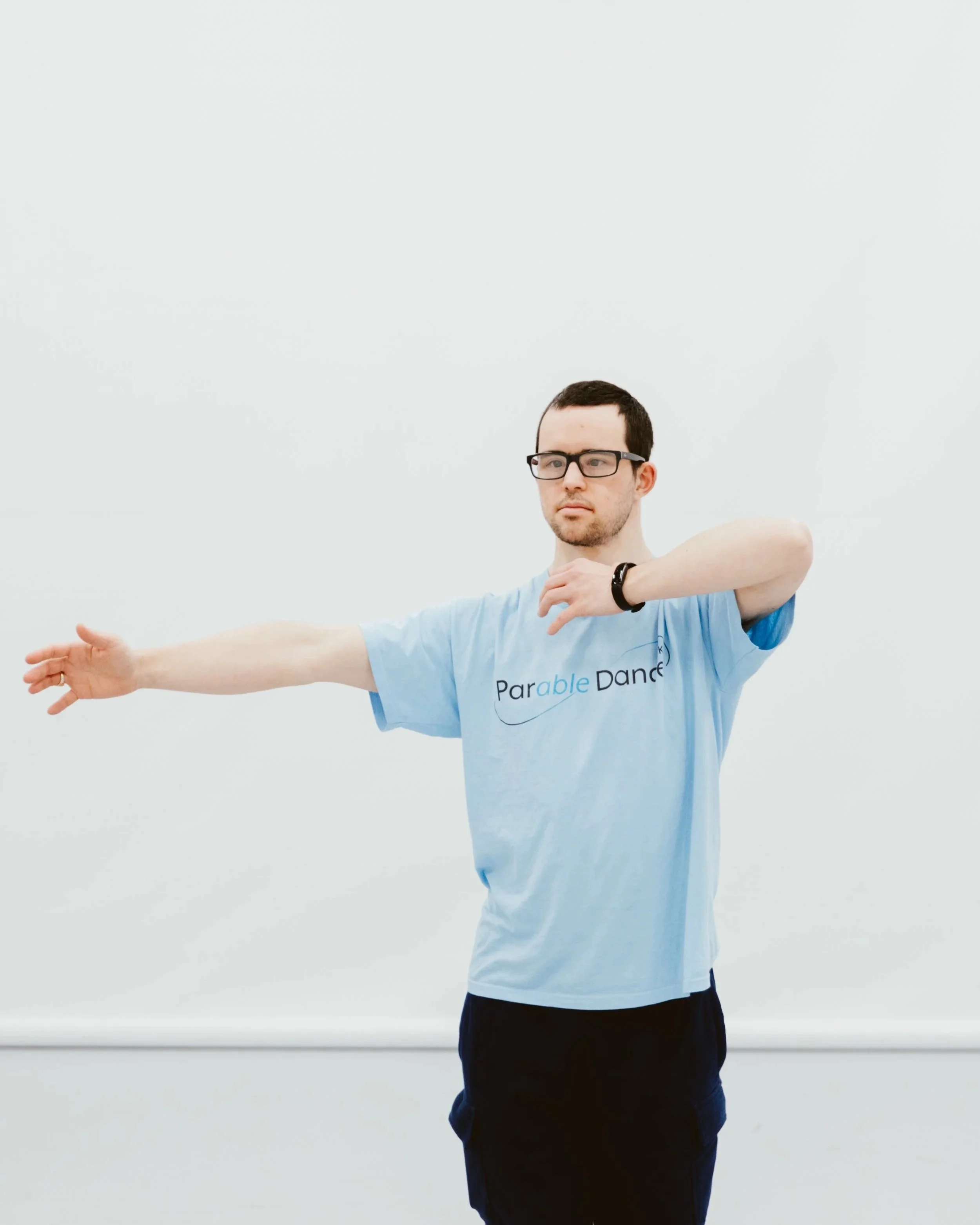 A man wearing glasses and a blue T-shirt and black trousers is standing with one arm extended and the other raised near his face, performing a dance move in a dance studio.