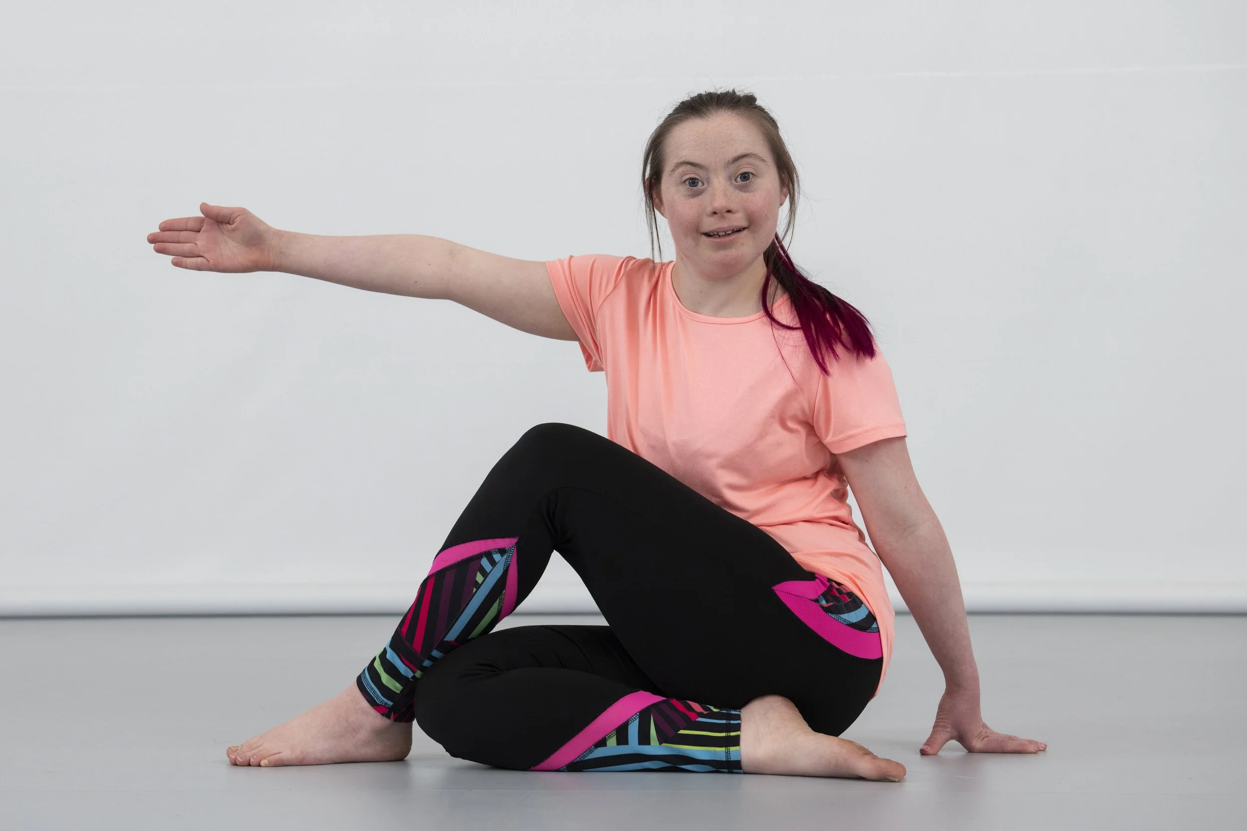 A young girl with hair tied back, wearing a peach-colored T-shirt, sitting on the floor in a yoga pose with her left hand on the ground, her right arm extended out to the side, and smiling at the camera.