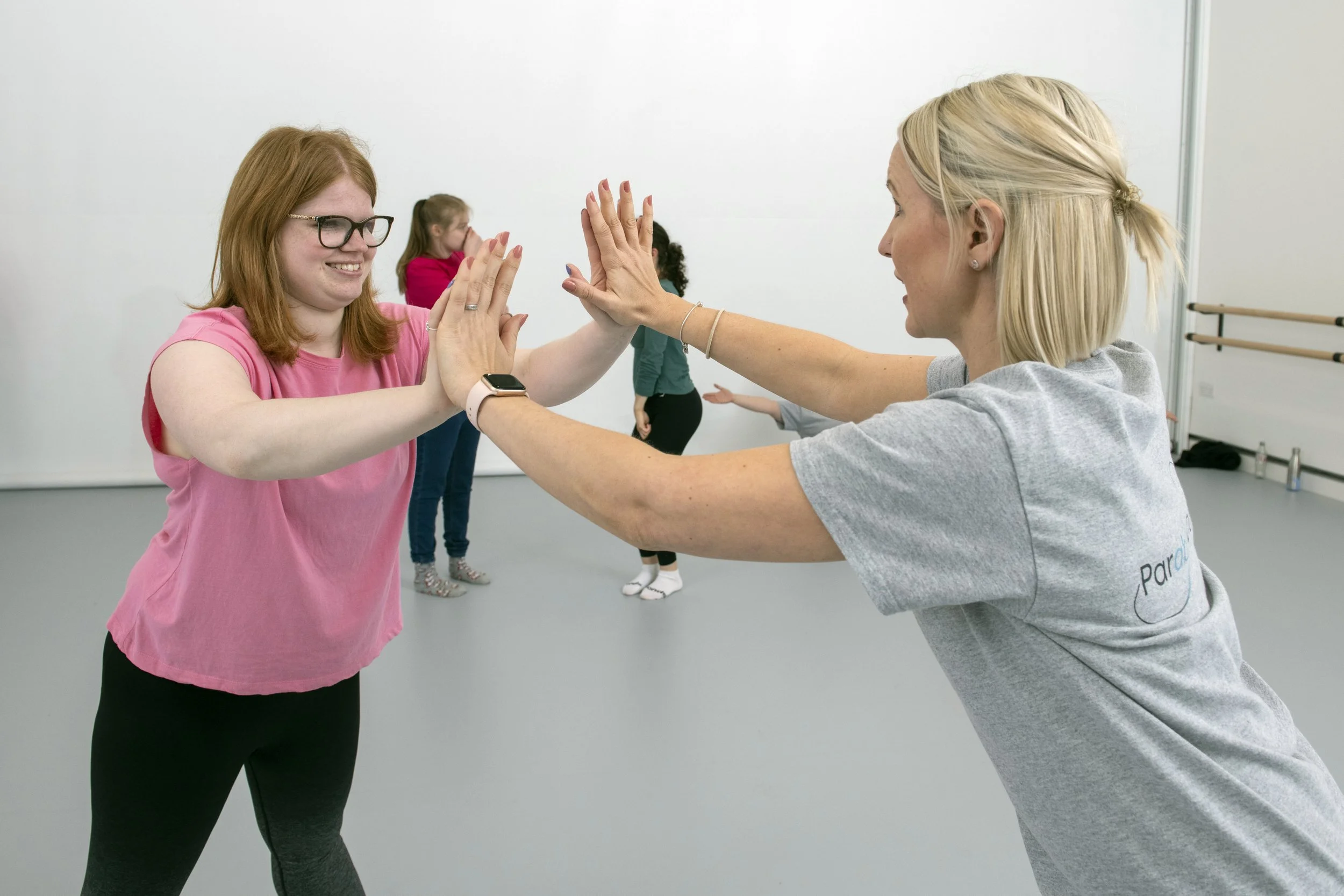 Women participating in a dance or exercise class high-fiving each other in a studio with a white wall and ballet bar in the background.